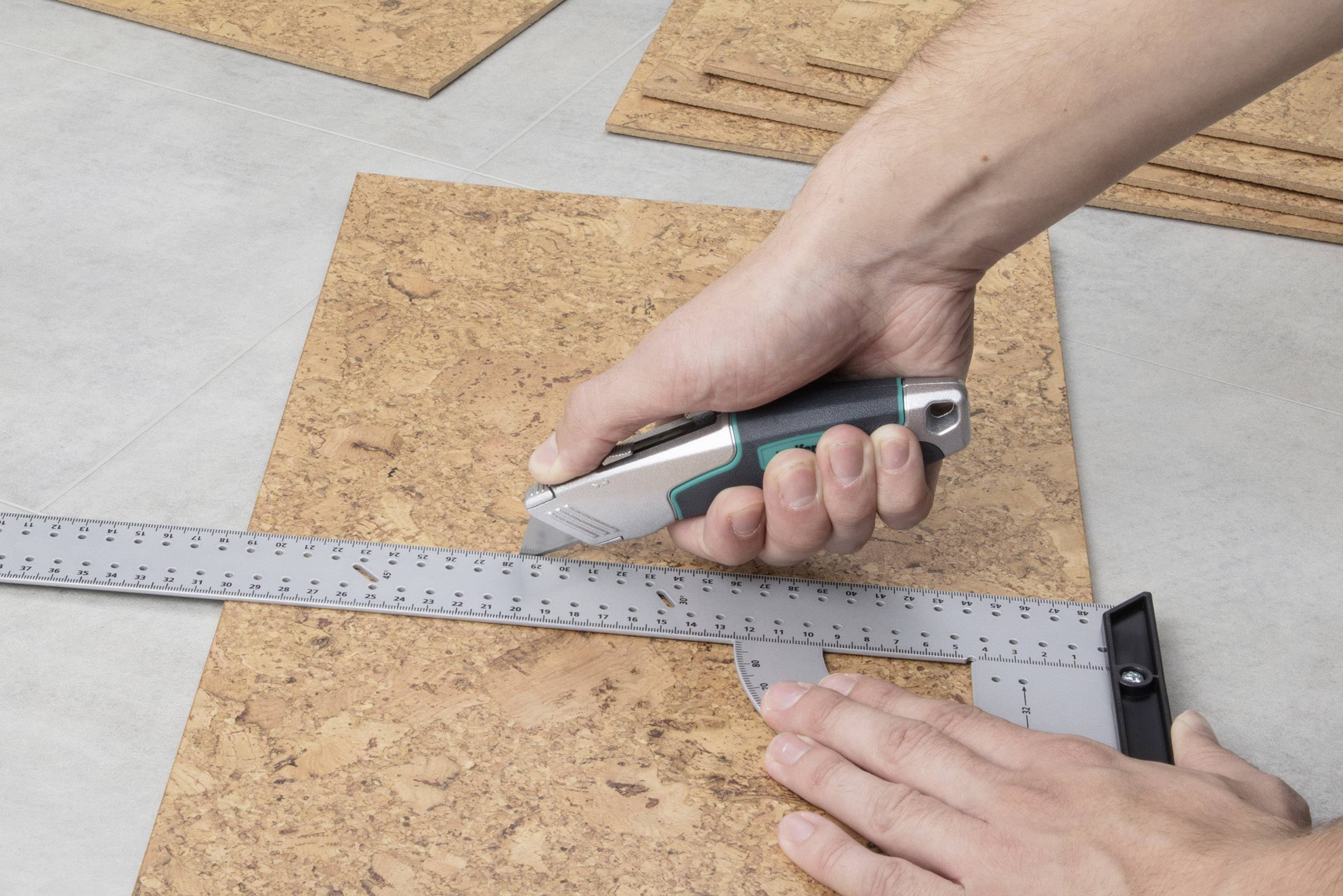 Eine Person schneidet ein Korkbrett mit einem Cuttermesser entlang eines Metalllineals. Hände halten das Messer und das Lineal fest.