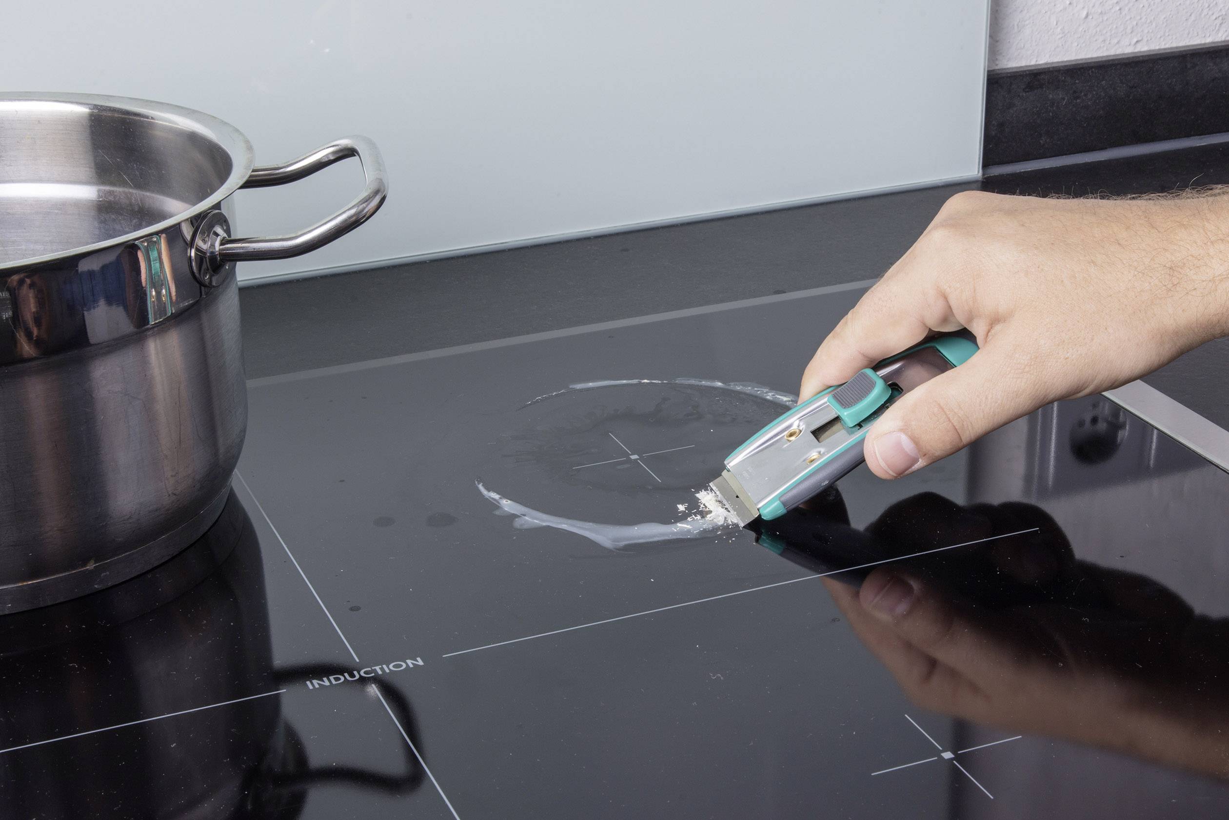 Eine Hand entfernt mit einem Schaber einen getrockneten Fleck von einem Induktionskochfeld. Links steht ein Edelstahltopf.