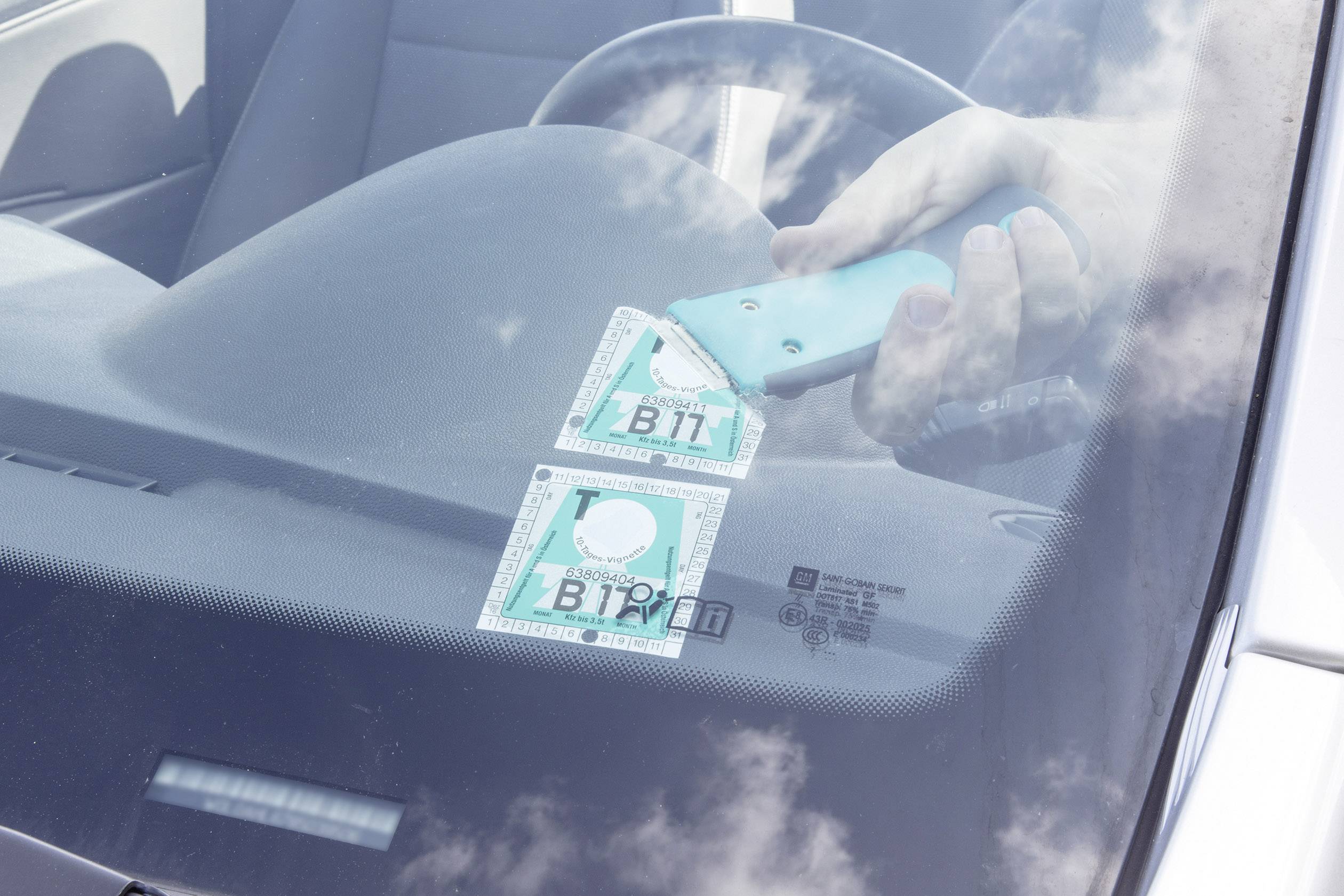 Ein Auto mit Autobahnvignette: Ein blauer Handabstreifer wischt zwei Vignetten von der Windschutzscheibe ab.
