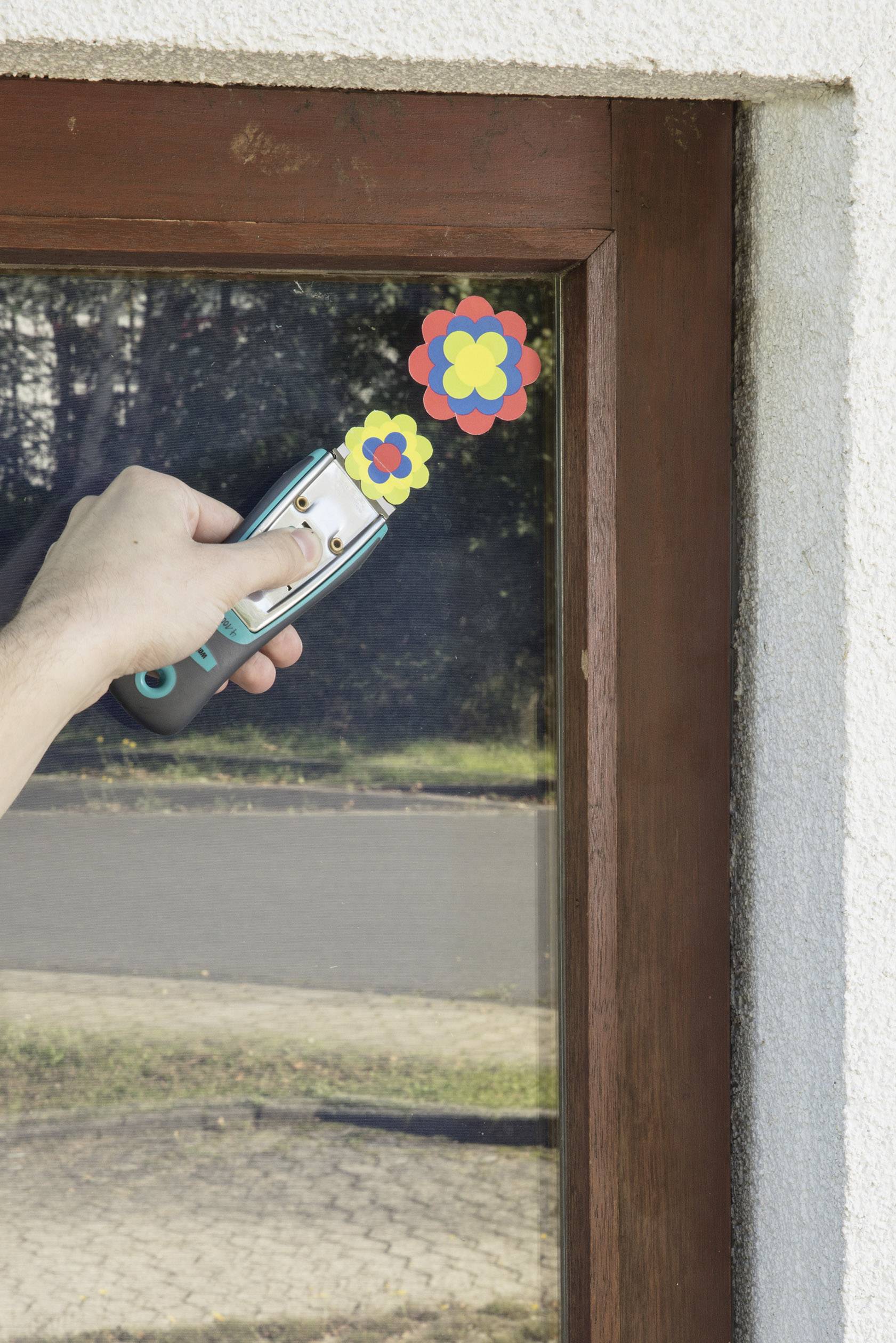 Hand entfernt mit Schaber bunte Blumenaufkleber von Fensterscheibe, draußen Straße und Bäume sichtbar.