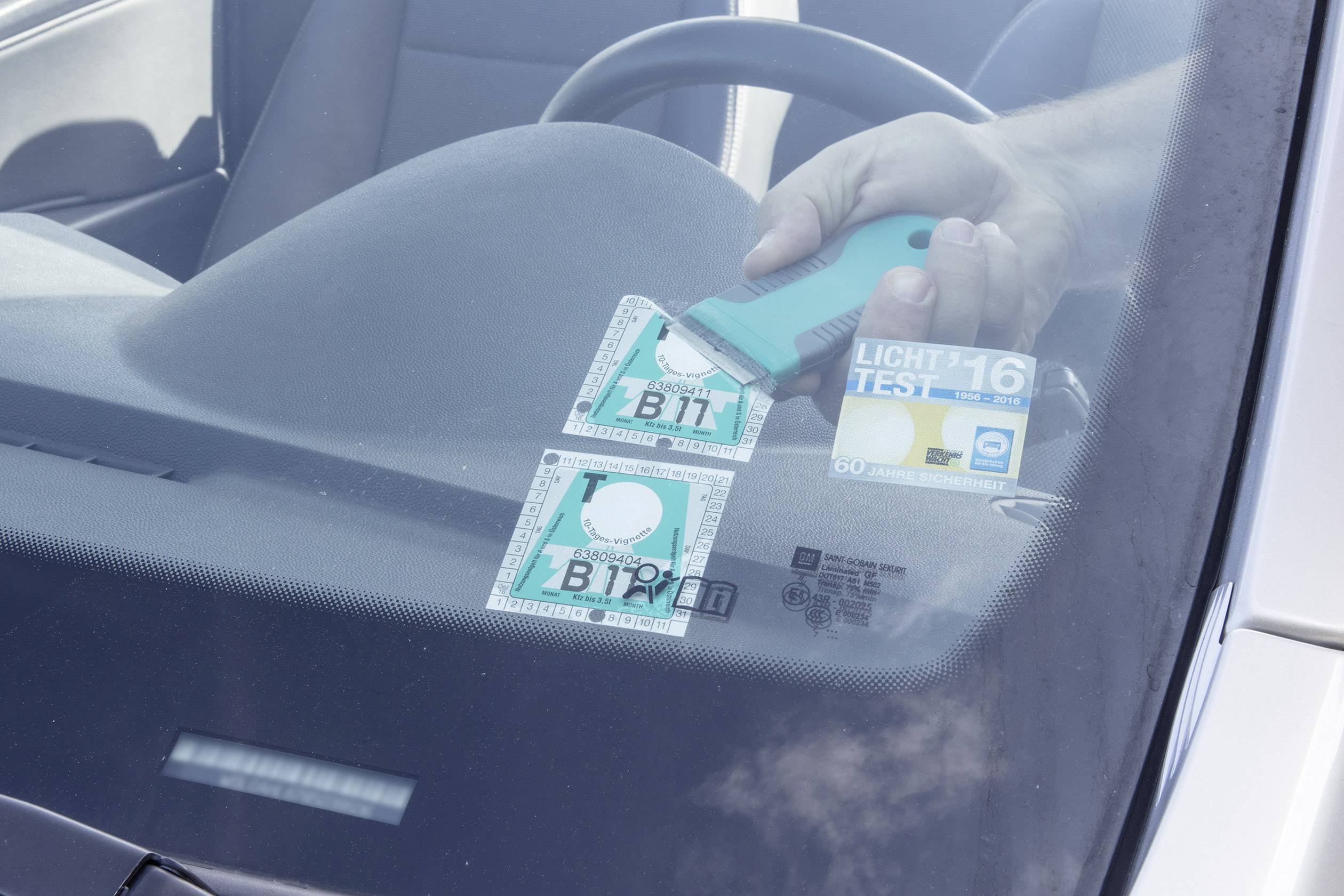 Eine Hand entfernt mit einem Werkzeug eine Umweltplakette von der Windschutzscheibe eines Autos.