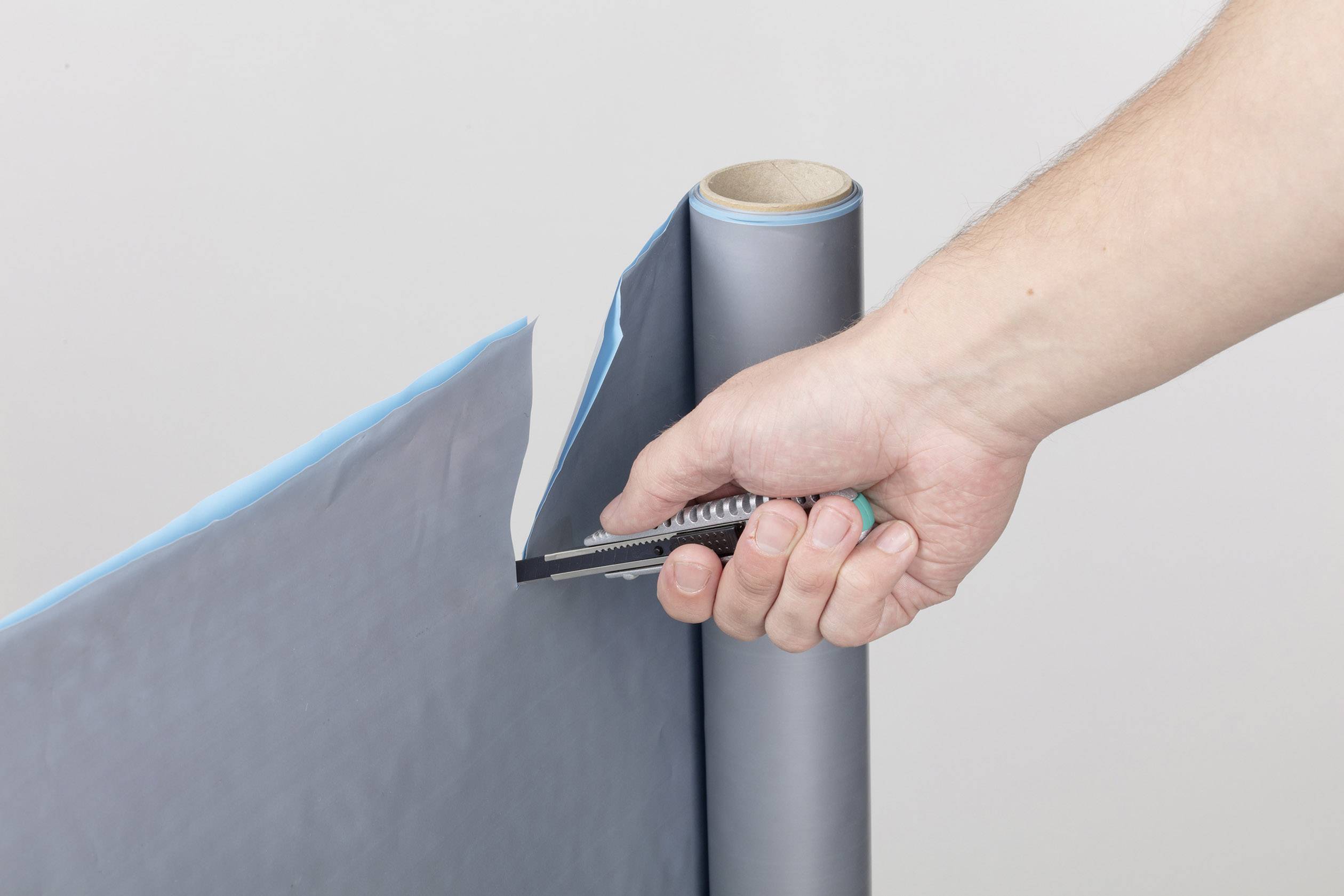 Person schneidet mit einem Teppichmesser ein dünnes, blaues Material von einer Rolle ab.