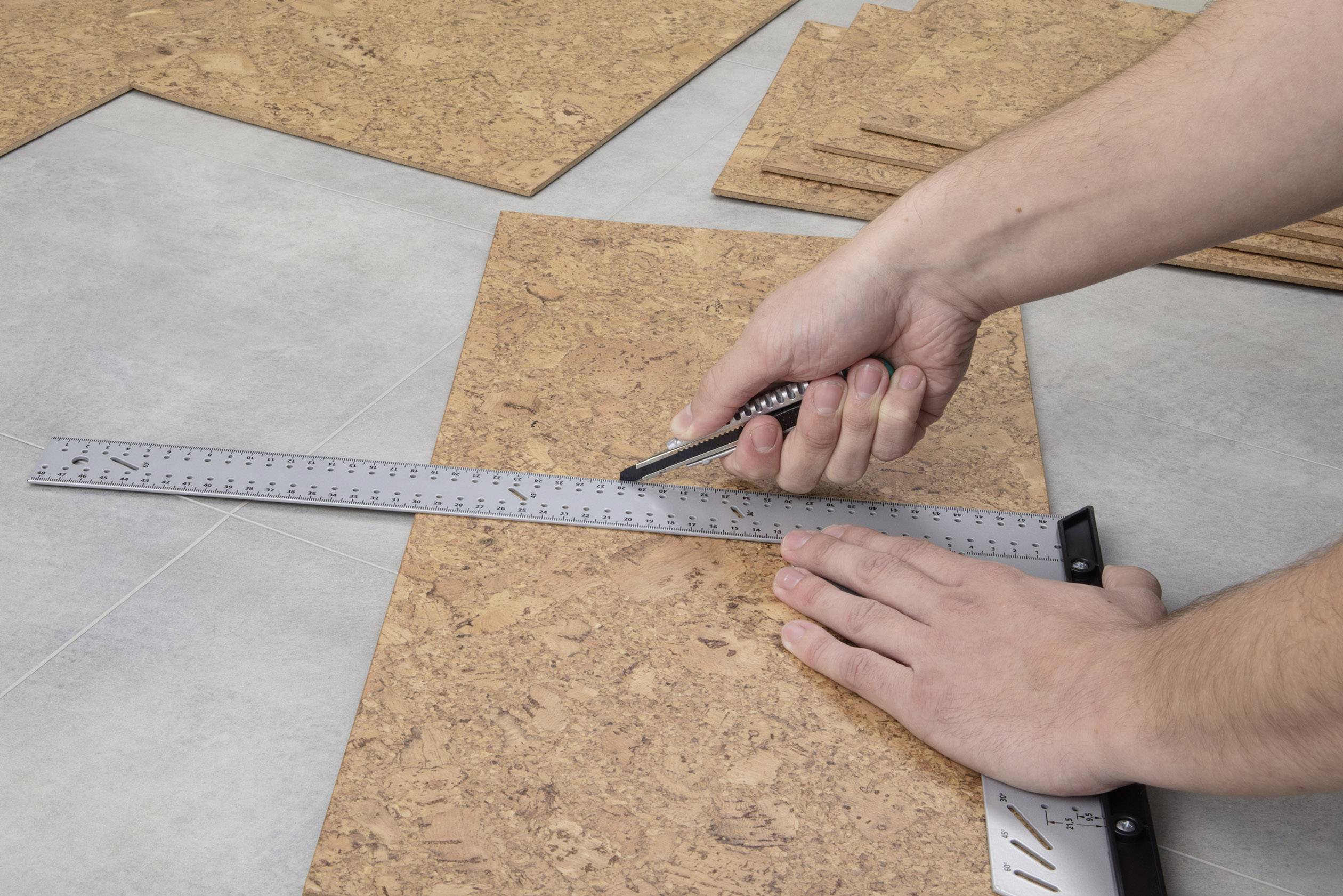 Eine Person schneidet eine Korkfliese mit einem Cuttermesser entlang eines Metall-Lineals auf einem grauen Boden.