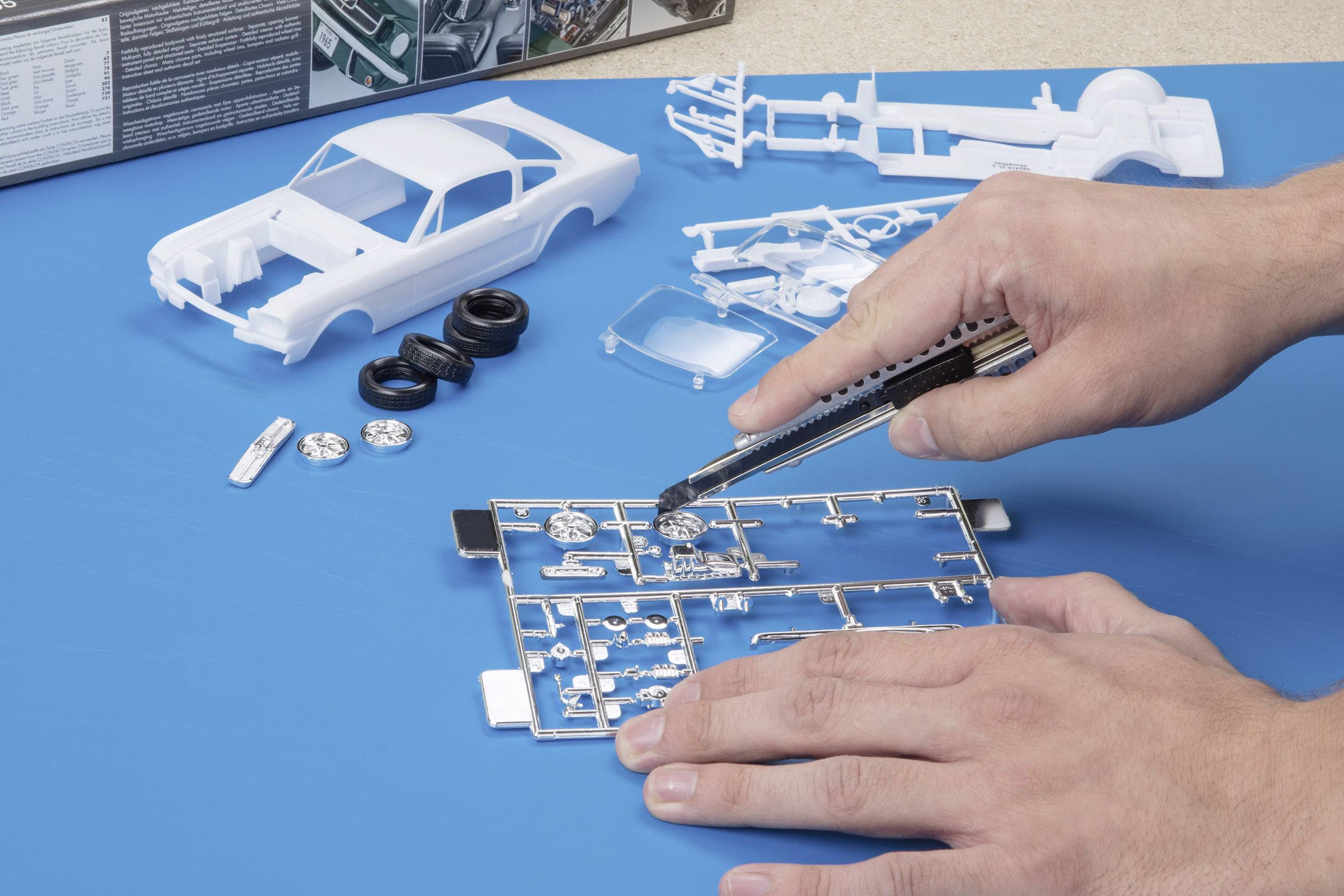 Ein Modellauto-Bausatz wird zusammengebaut. Hände schneiden Teile aus einem Plastikrahmen. Im Hintergrund befinden sich weitere Bauteile.