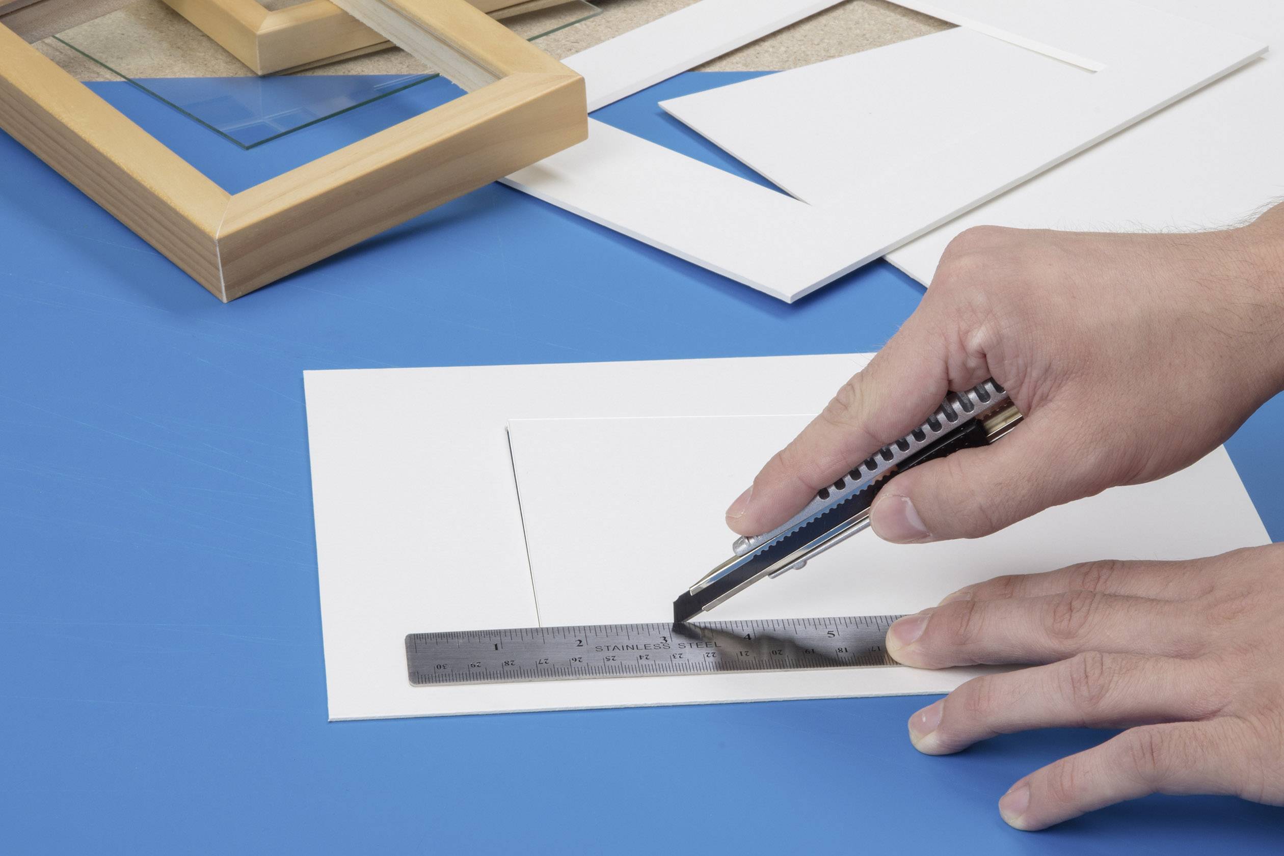 Eine Hand schneidet mit einem Cuttermesser einen rechteckigen Ausschnitt in weißes Papier auf einer blauen Unterlage, daneben ein Lineal und Rahmen.
