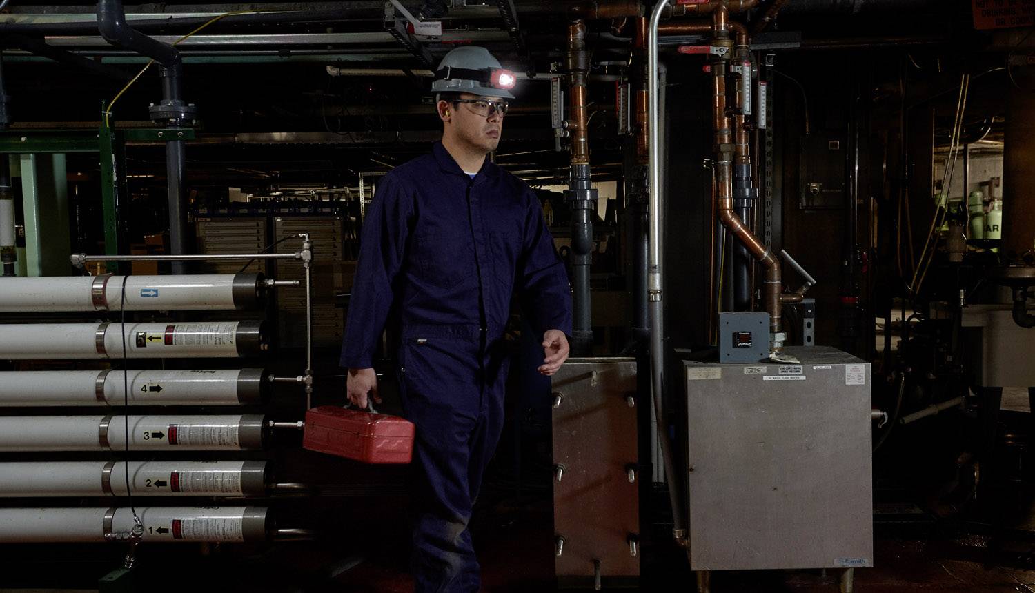 Ein Arbeiter in blauer Schutzkleidung und Helm trägt einen roten Werkzeugkasten in einem industriellen Raum mit Rohren und Maschinen.