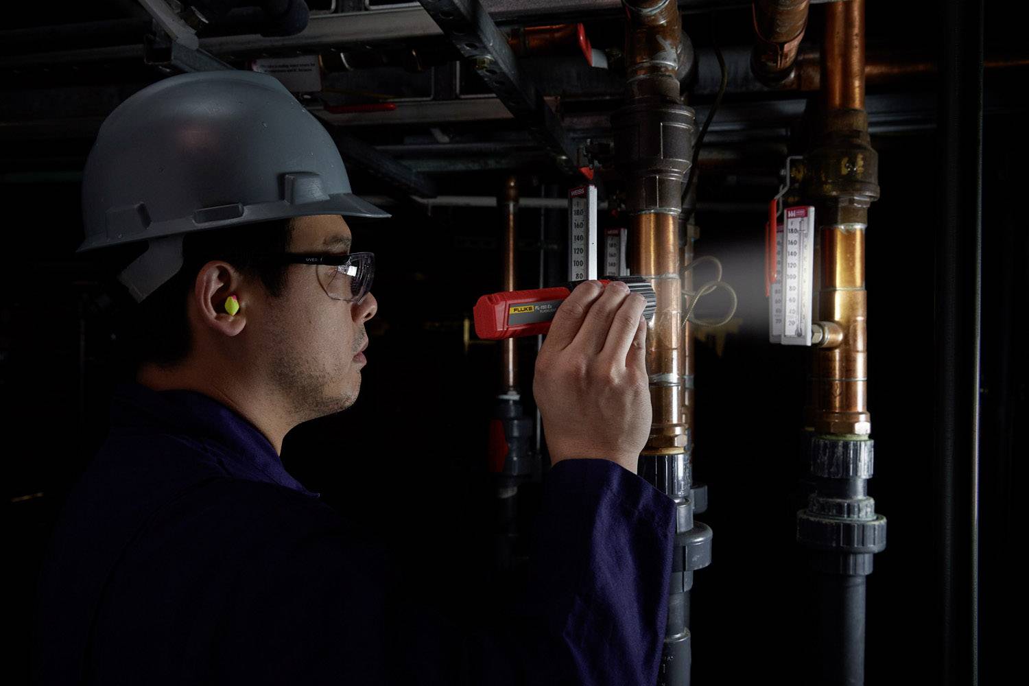Eine Person mit Schutzhelm und Brille inspiziert Rohre in einem industriellen Umfeld mit einem roten Gerät zur Prüfung von Gaslecks.