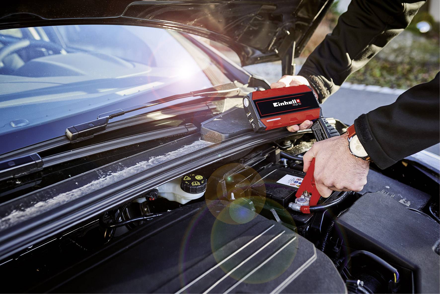 Person montiert eine Autobatterie mit einem 'Einhell'-Gerät im Motorraum eines Autos.