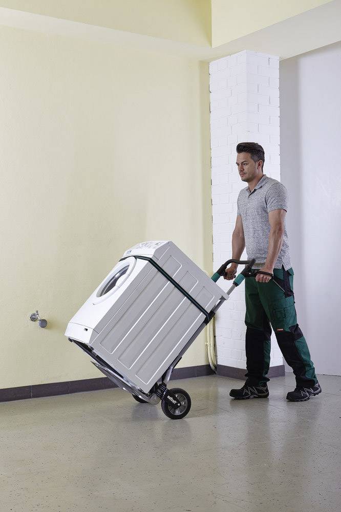 Ein Mann in Arbeitskleidung transportiert eine Waschmaschine mit einem Sackkarren. Er bewegt sich in einem Raum mit hellen Wänden.