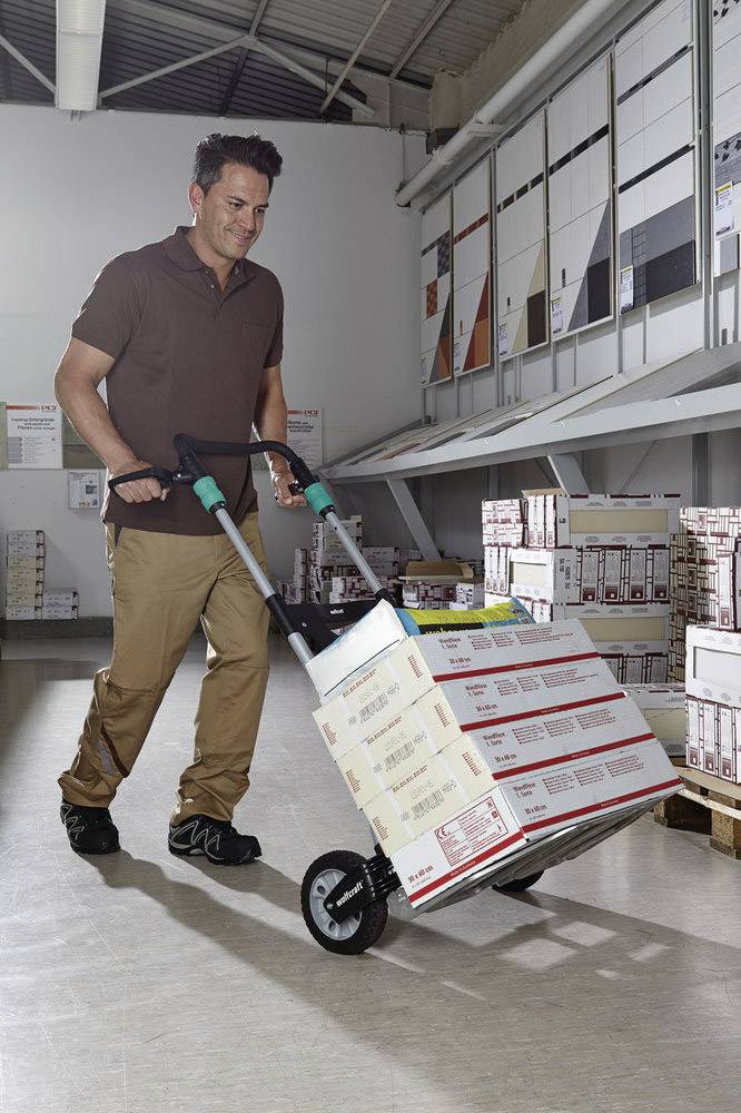 Ein Mann transportiert mit einem Handwagen mehrere gestapelte Kartons durch ein Lager. Kartons sind rechteckig und beschriftet.