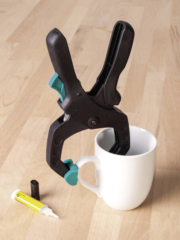 Eine schwarze Klemme hält eine weiße Tasse aufrecht. Daneben liegt ein offener Klebstofftube auf einem Holztisch.