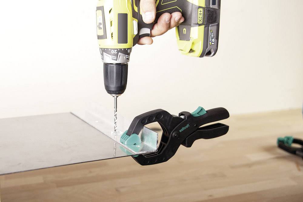 Eine Person bohrt mit einer gelb-schwarzen Bohrmaschine in eine transparente Platte, die mit einer grünen Klemme fixiert ist.