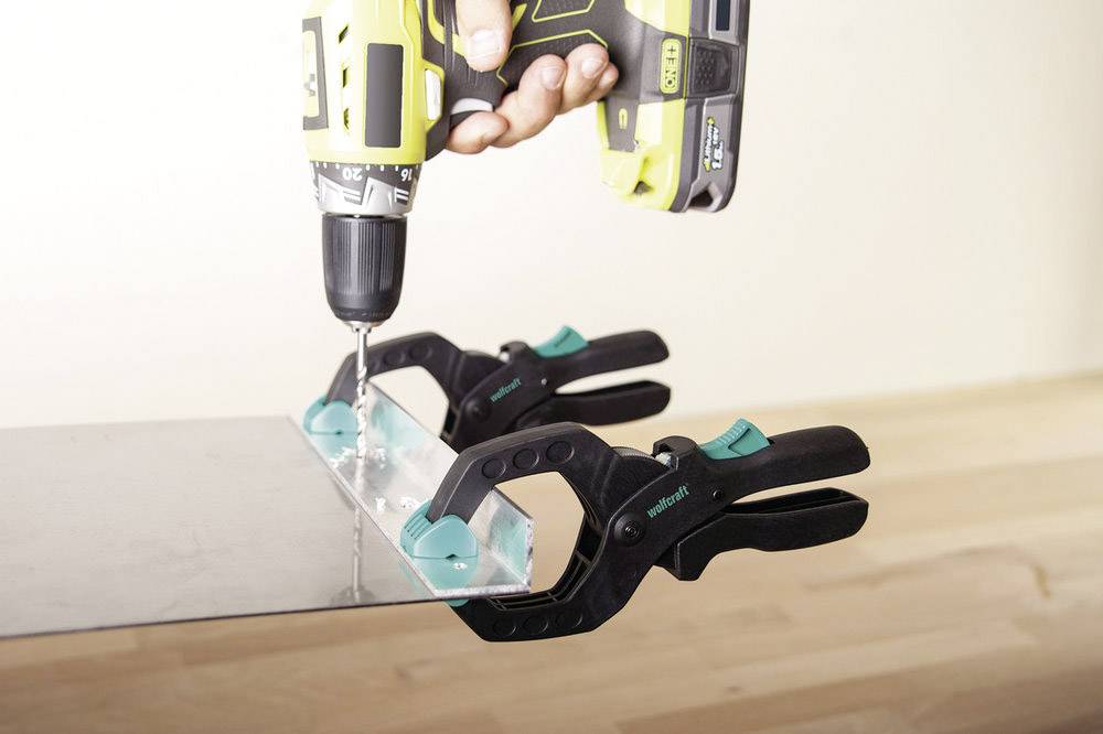 Eine Person bohrt mit einem Akku-Bohrschrauber Löcher in eine Metallplatte, die mit zwei Klemmen auf einer Holzfläche fixiert ist.