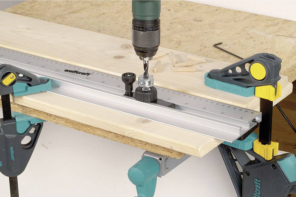 Ein Holzstück wird mithilfe von Klemmen auf einem Tisch fixiert. Eine Bohrmaschine ist an einem Lineal befestigt, um präzise Bohrungen durchzuführen.
