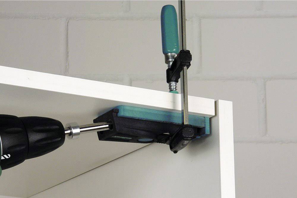 Ein Bohrer bringt Löcher in ein weißes Regalbrett, das mit einer Schraubzwinge an einer Lehre befestigt ist.