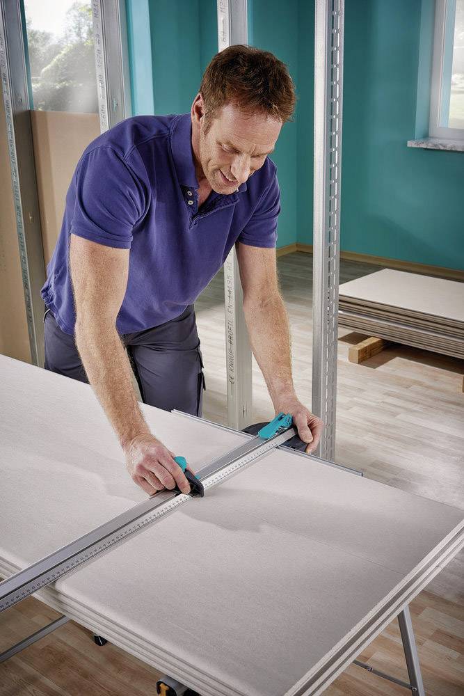 Ein Mann misst mit einem Lineal und Schneidegerät auf einer Trockenbauplatte in einem Raum, der im Bau oder Renovierung ist.