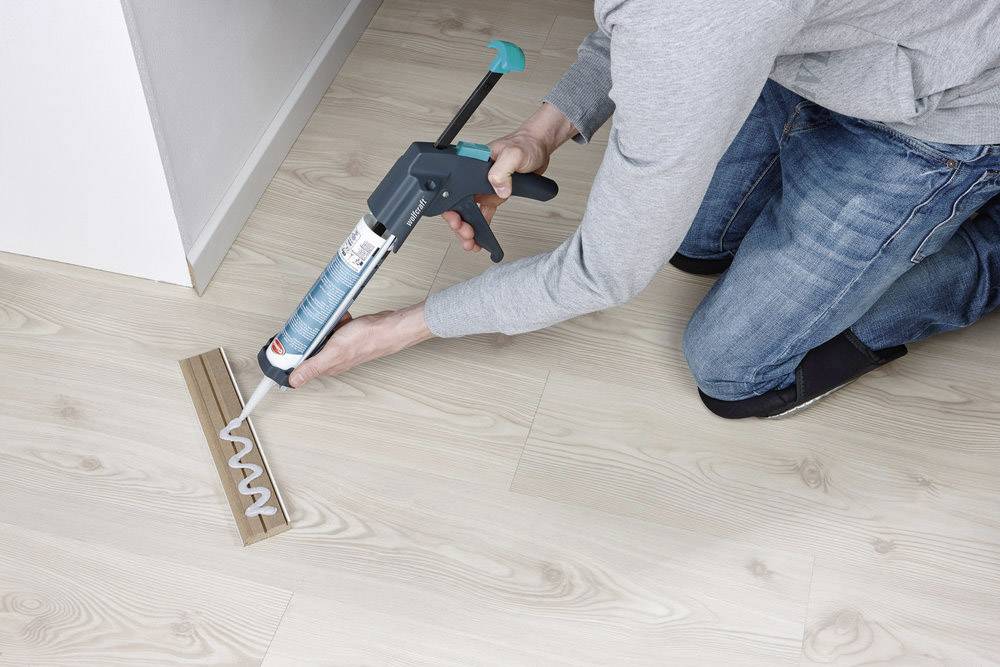 Person kniet auf Holzboden und trägt mit einer Kartuschenpistole Klebstoff in einer Zickzacklinie auf ein Holzstück auf.