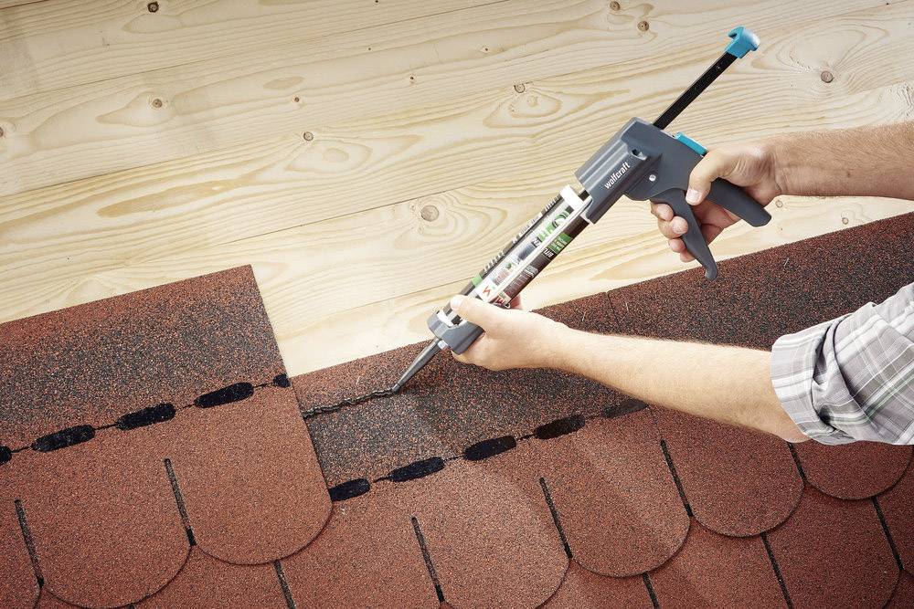 Person verwendet eine Kartuschenpistole, um Dachziegel zu versiegeln. Ziegel sind rot und Dach ist aus Holz.