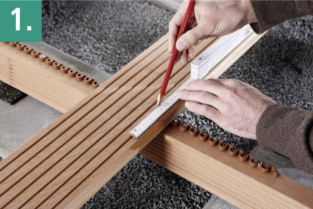 Handwerkliche Aufgabe: Zwei Hände messen und markieren präzise die Position auf einer Holzlatte mit einem roten Stift und einem Metermaß.