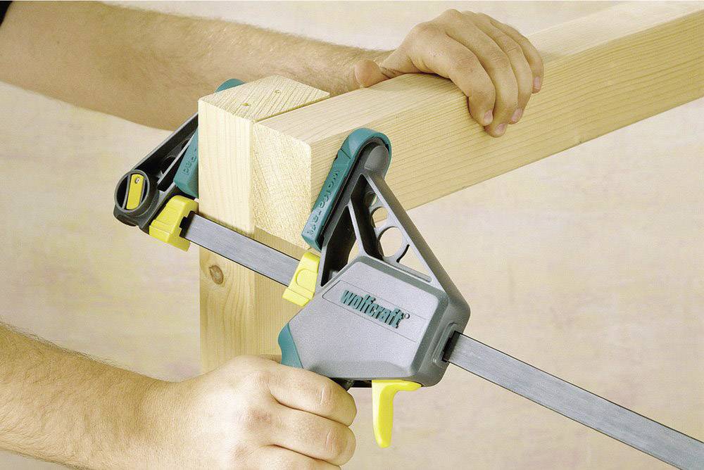 Jemand verwendet eine Holz-Zwinge, um zwei Holzbretter im rechten Winkel zu verbinden. Die Zwinge ist grau und gelb.