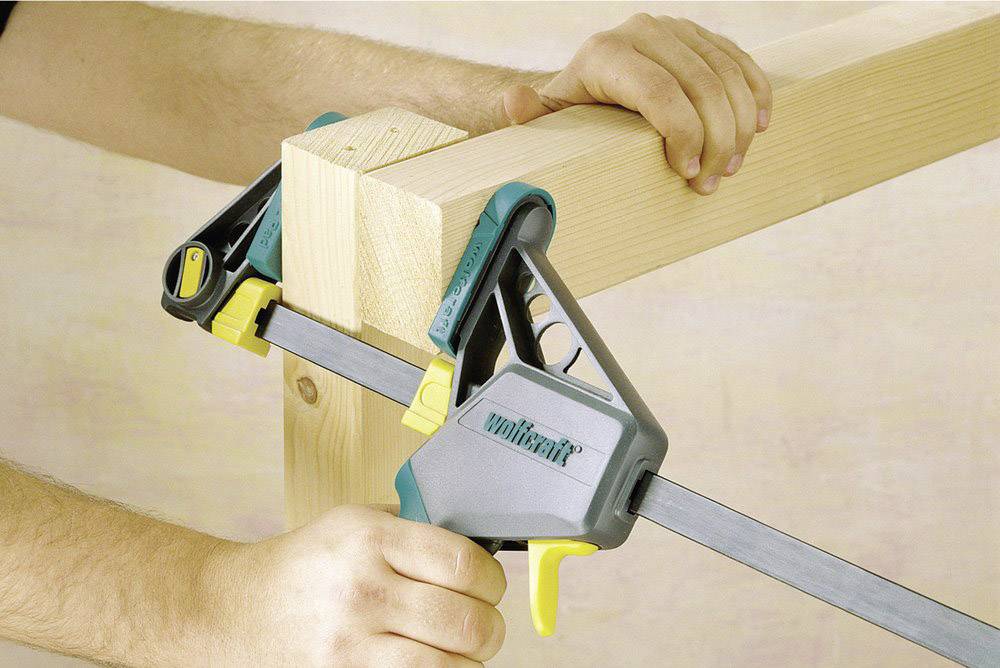Eine Person benutzt eine Einhandzwinge, um zwei Holzstücke in einem rechten Winkel zusammenzuhalten. Die Klemme hat einen gelben Griff.