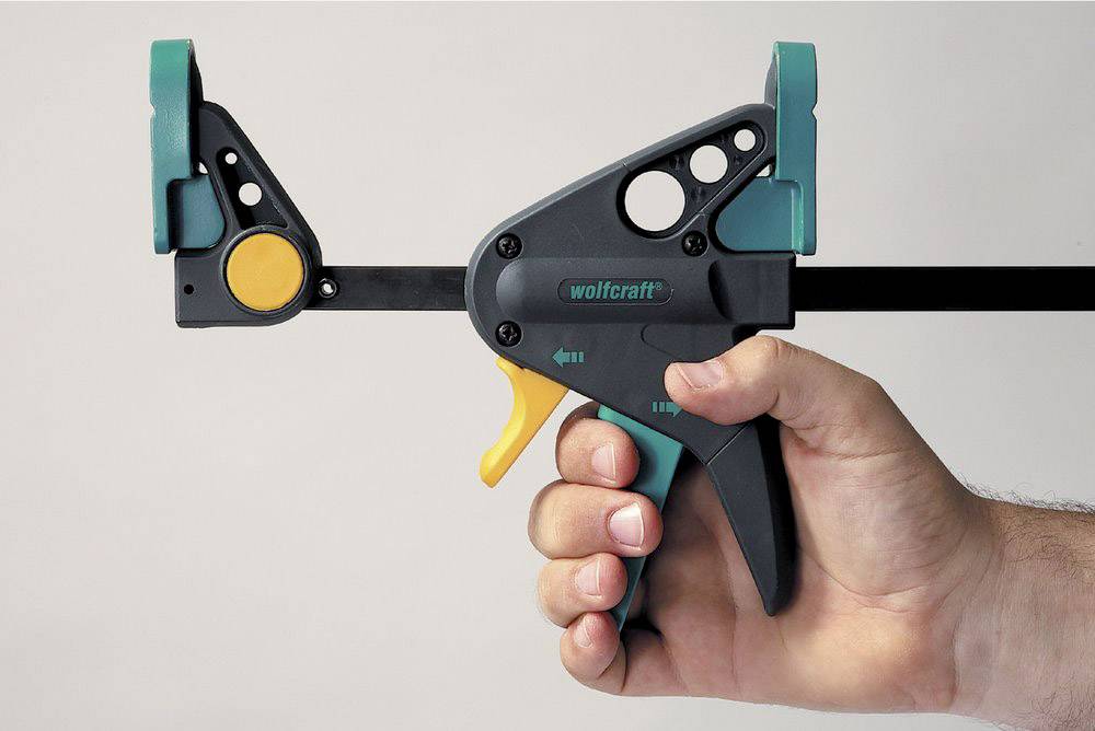 Eine Hand hält eine grüne und schwarze Einhandzwinge mit gelbem Hebel von Wolfcraft. Die Zwinge ist geöffnet und einsatzbereit.
