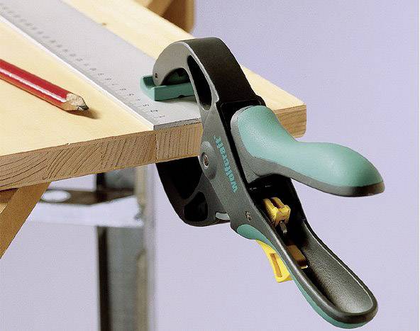 Eine Schraubzwinge hält ein Holzbrett sicher auf einem Tisch fest. Daneben liegt ein Bleistift und ein Metalllineal.