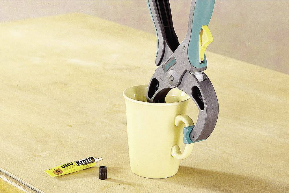 Eine Schraubzwinge hält eine gelbe Tasse auf einem Holztisch fest. Daneben liegt eine Tube Uhu-Kleber ohne Verschluss.