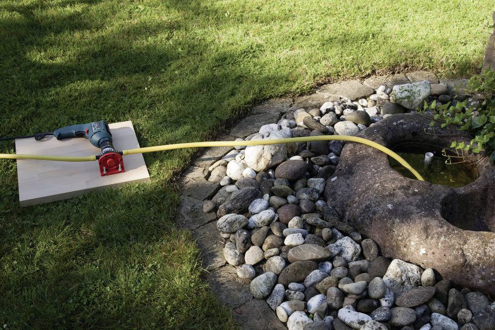 Bohrmaschine mit Schlauch pumpt Wasser in einen Steinbrunnen, umgeben von Kieselsteinen und Gras.