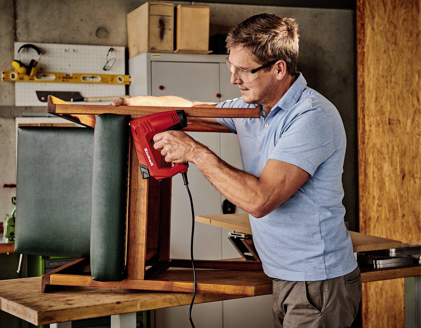 Ein Mann in einer Werkstatt tackert neues Polster auf einen umgedrehten Stuhl. Er trägt eine Schutzbrille und benutzt einen roten Tacker.
