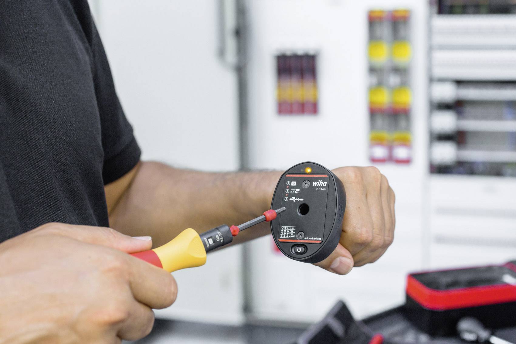 Eine Person hält ein elektrisches Prüfgerät und testet eine Spannung. Im Hintergrund sind unscharfe Schaltschränke zu sehen.