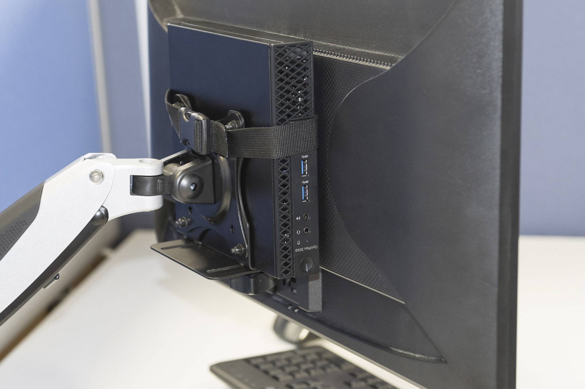 Ein kleiner Computer ist mit einem Band an der Rückseite eines Monitors befestigt, der an einem beweglichen Monitorarm montiert ist.
