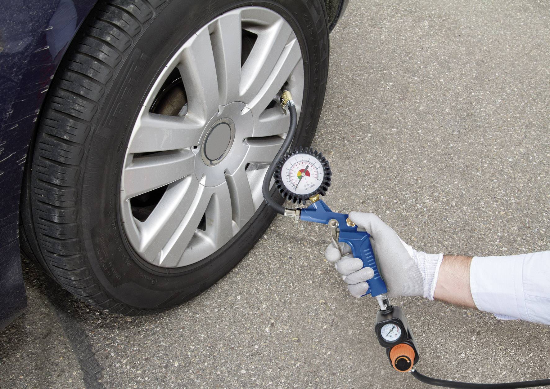 Eine Person misst den Reifendruck an einem Autoreifen mit einem Manometer. Der Reifen ist auf einem Asphaltuntergrund.