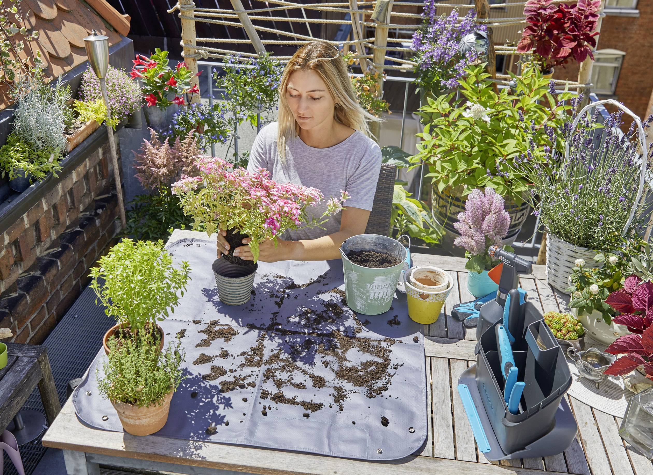 Eine Frau pflanzt Blumen auf einem Balkon voller grüner Pflanzen und bunter Blumen in Töpfen. Sie bereitet die Erde auf einem Tisch vor.