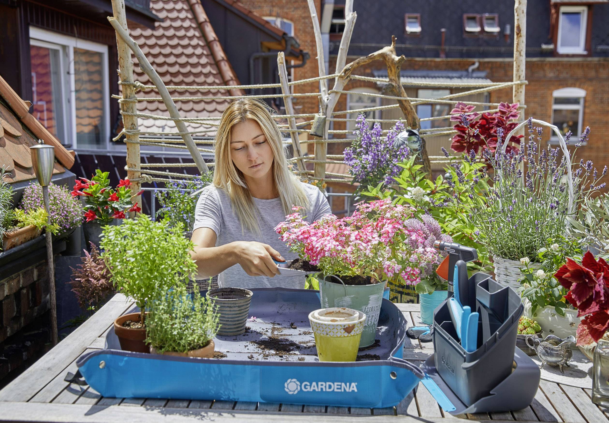 Eine Frau sitzt an einem Tisch auf einer Terrasse und pflanzt Blumen in Töpfe umgeben von blühenden Pflanzen und Gartenutensilien.