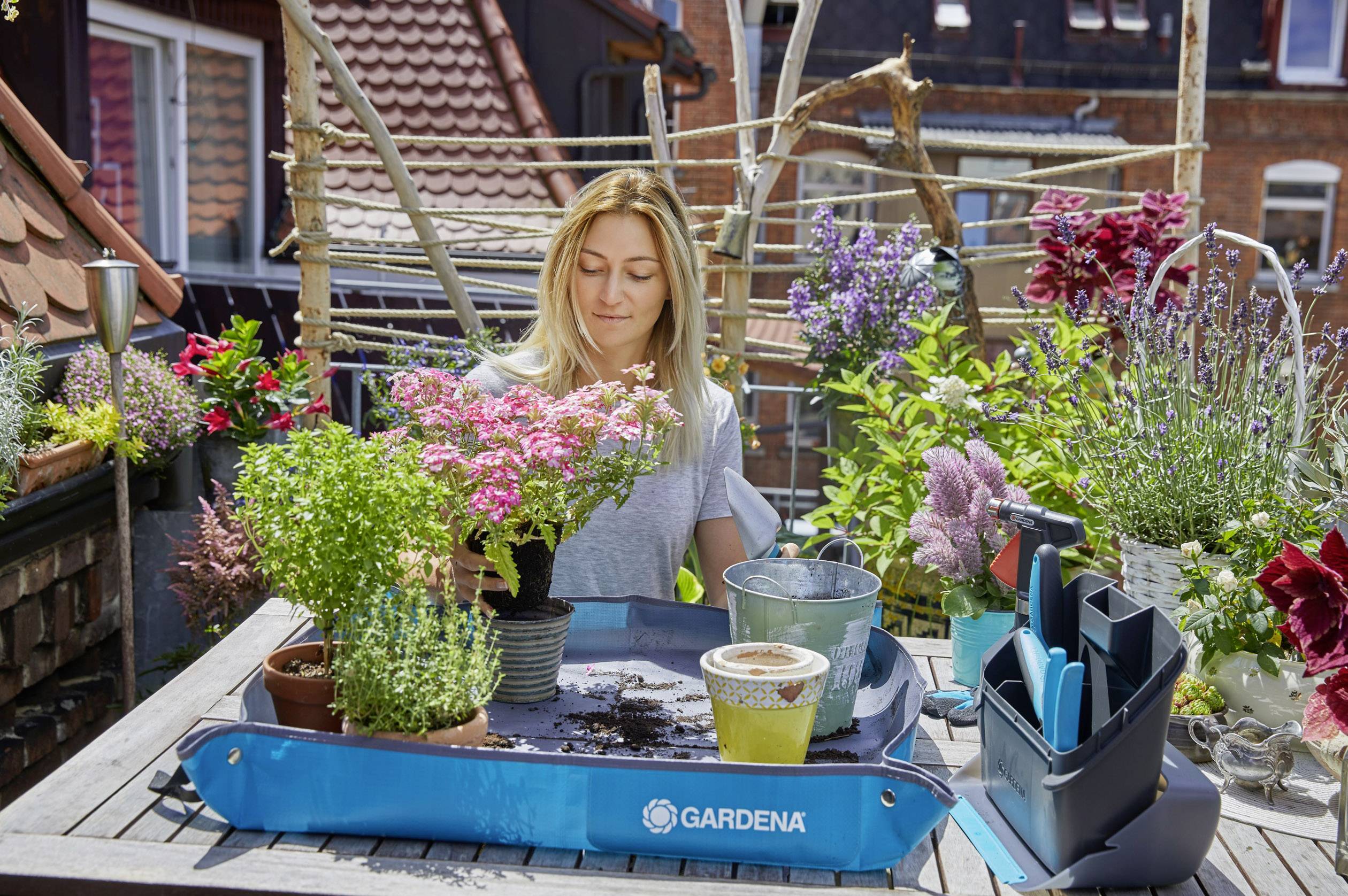 Frau topft Pflanzen auf einem Balkon um, umgeben von blühenden Blumen. Eine blaue Box mit Gartenwerkzeugen steht auf dem Tisch.