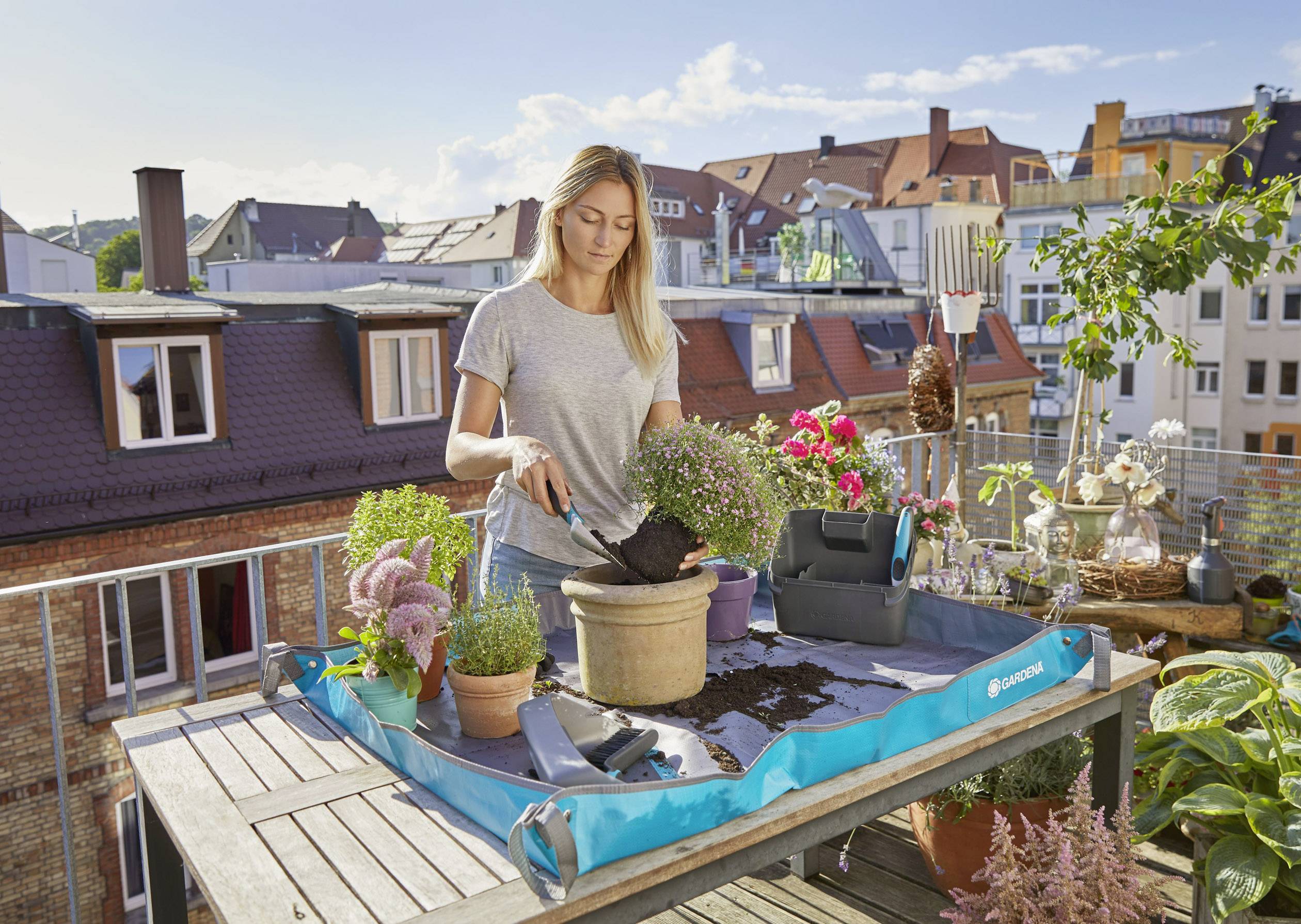 Eine Frau pflanzt Blumen auf einer Terrasse, umgeben von städtischen Gebäuden. Gartengeräte und Topfpflanzen sind auf einem Tisch verteilt.