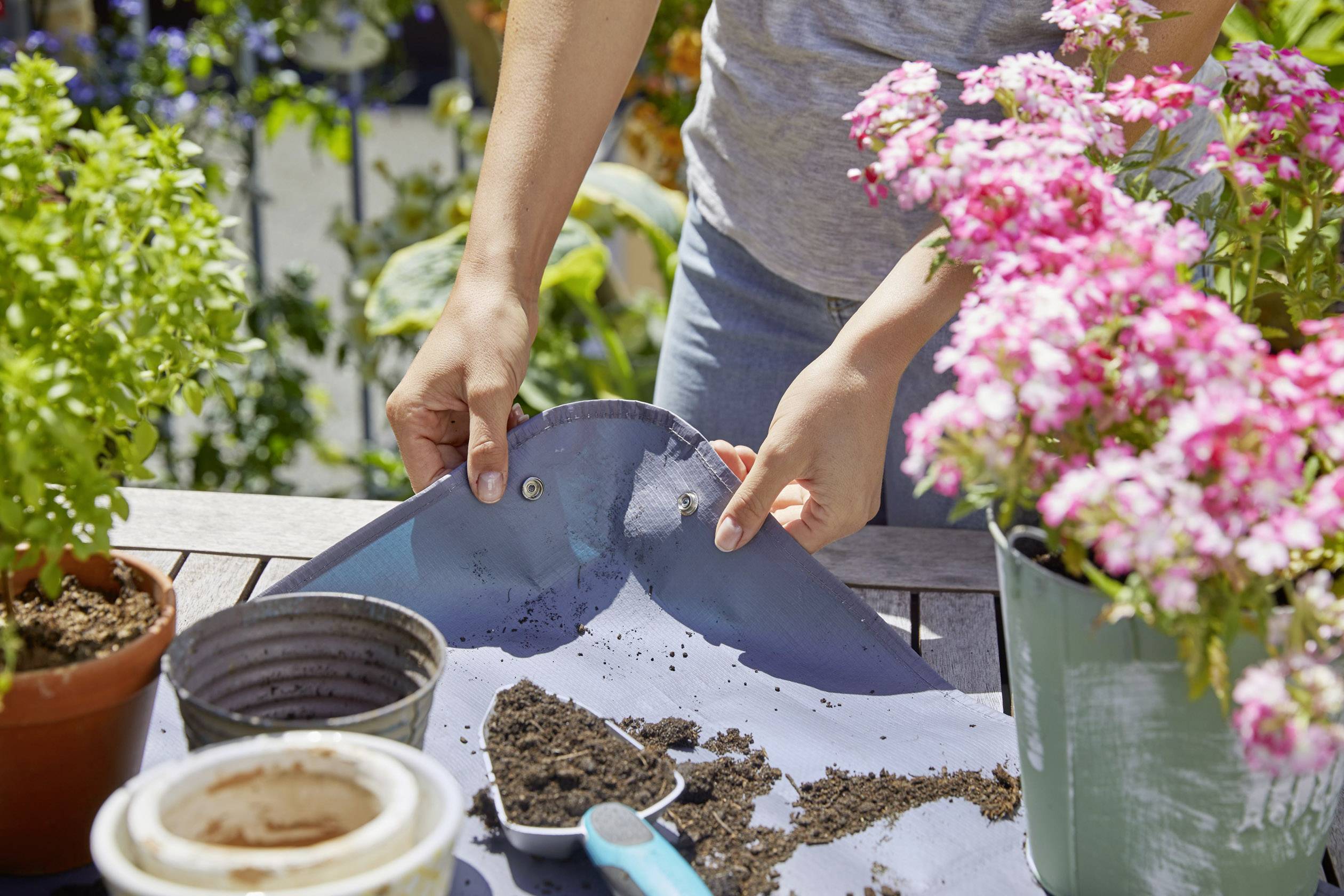 Person pflanzt Blumen in Töpfen auf einem Tisch im Freien. Erde, eine Schaufel und blühende Pflanzen sind sichtbar.
