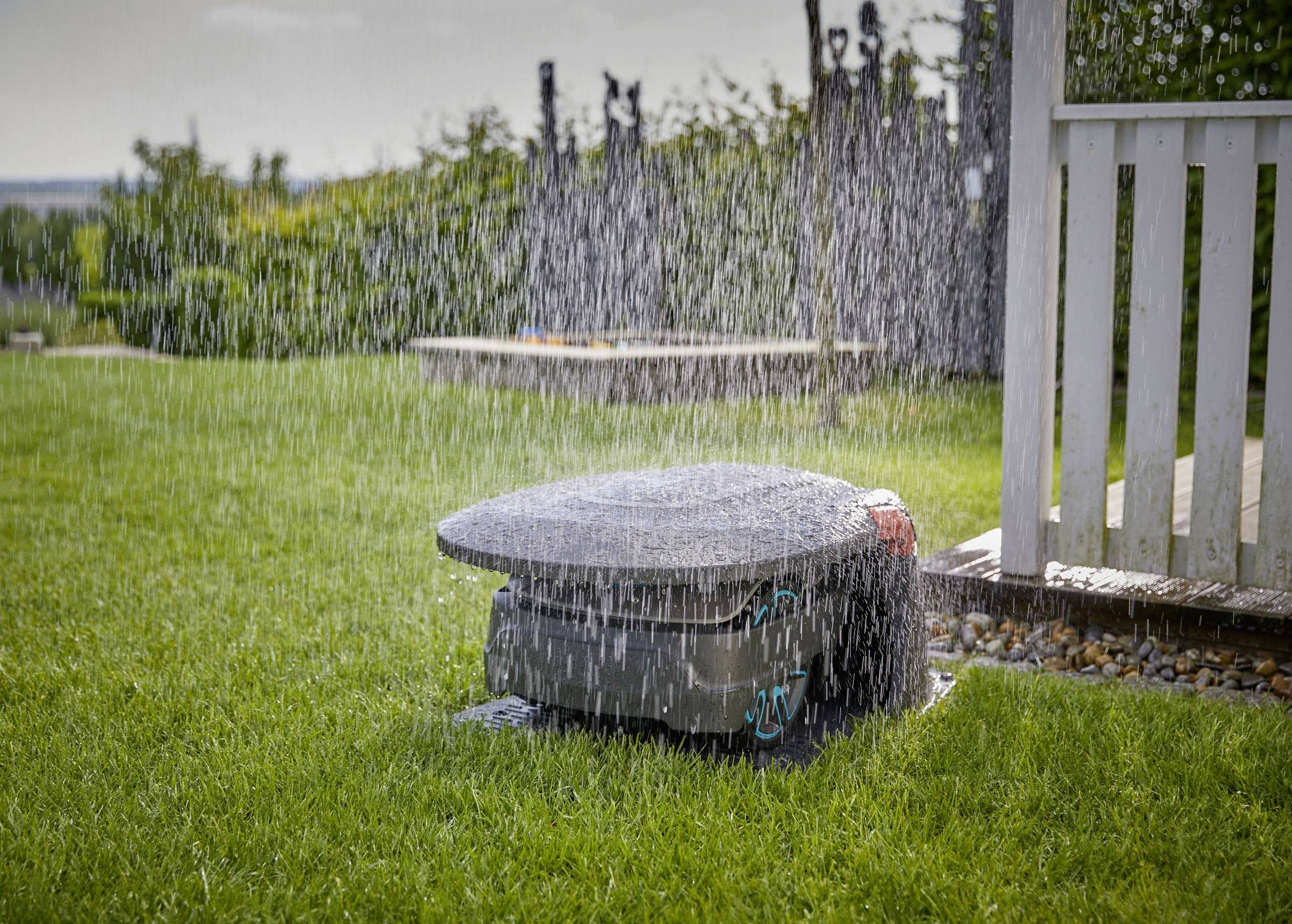 Ein Rasensprenger bewässert einen grünen Garten. Ein weißer Zaun ist im Hintergrund sichtbar. Das Wasser bildet einen gleichmäßigen Regen.