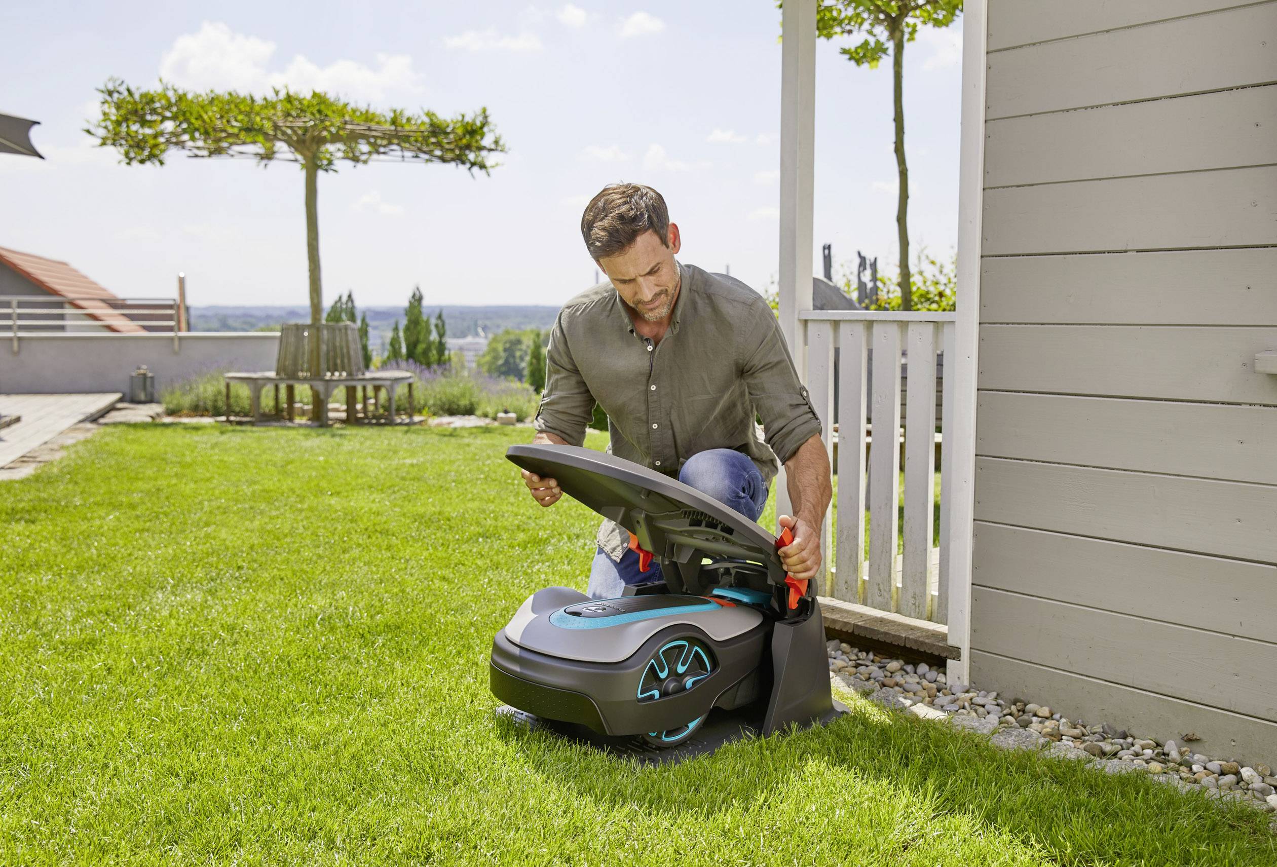 Ein Mann kniet auf einem Rasen und überprüft einen automatischen Rasenmäher neben einem Haus. Im Hintergrund ist ein Garten zu sehen.