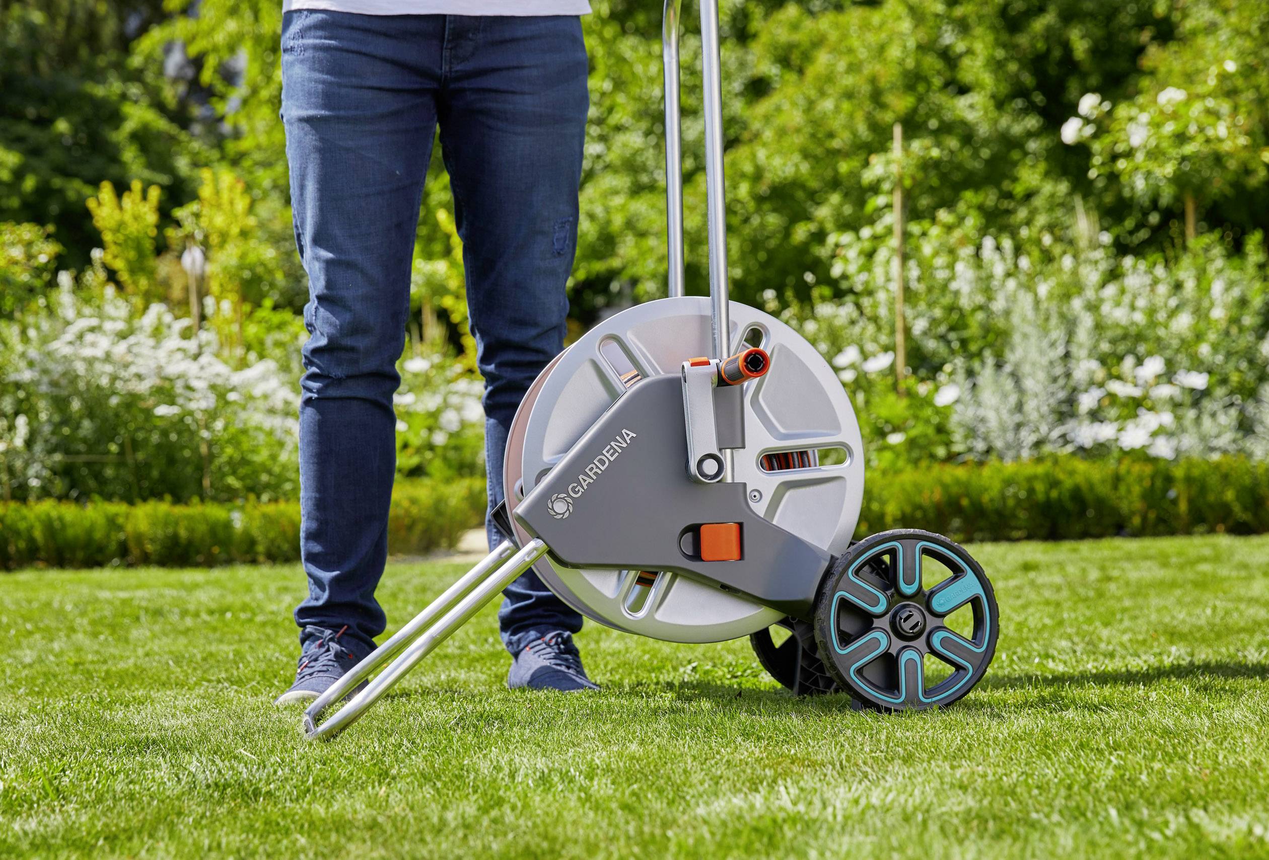 Person steht auf einem Rasen neben einem Gartenschlauchwagen. Hintergrund zeigt blühende Pflanzen.