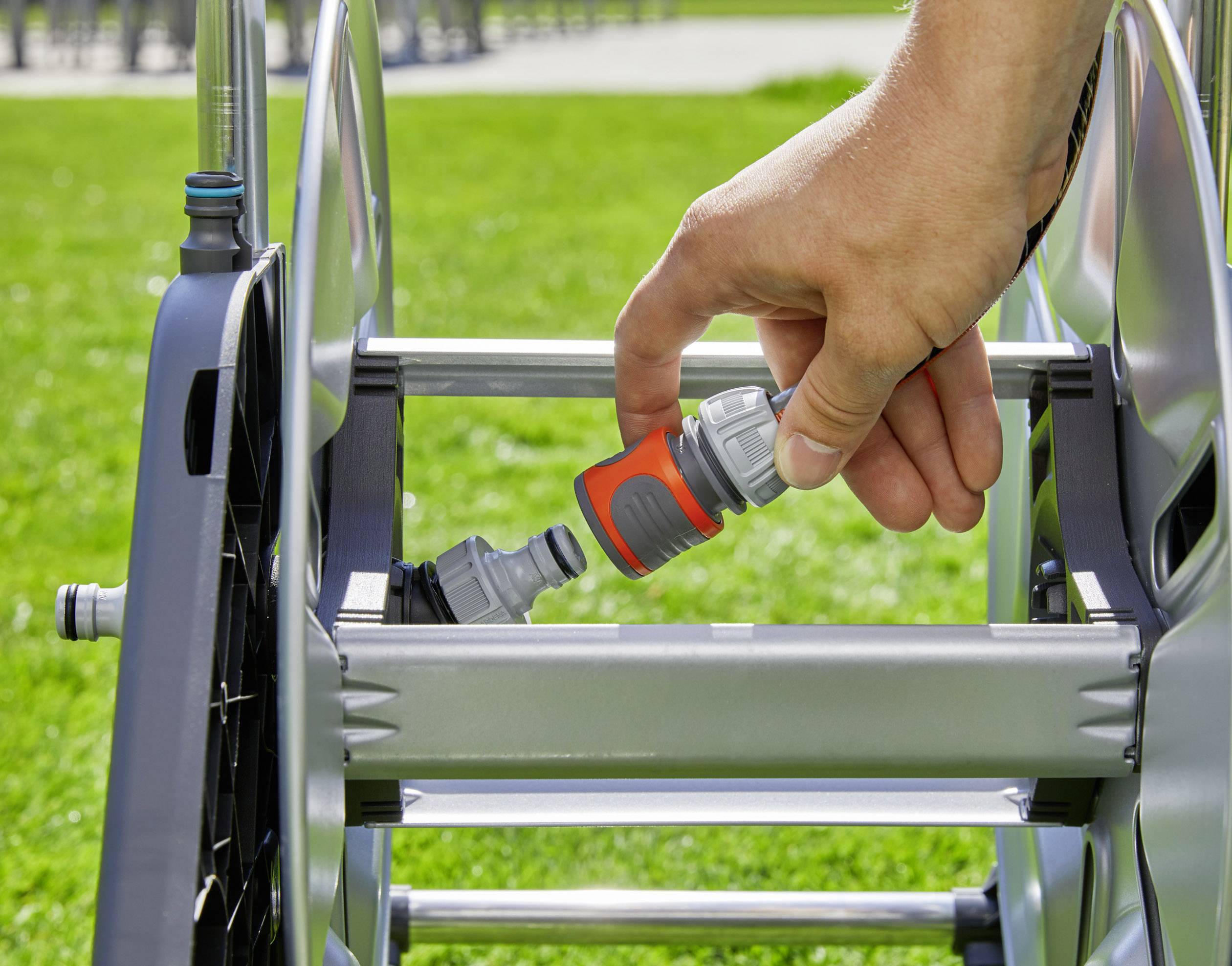 Eine Hand steckt einen Schlauchanschluss an einen Schlauchwagen im Garten an. Gras ist im Hintergrund sichtbar.