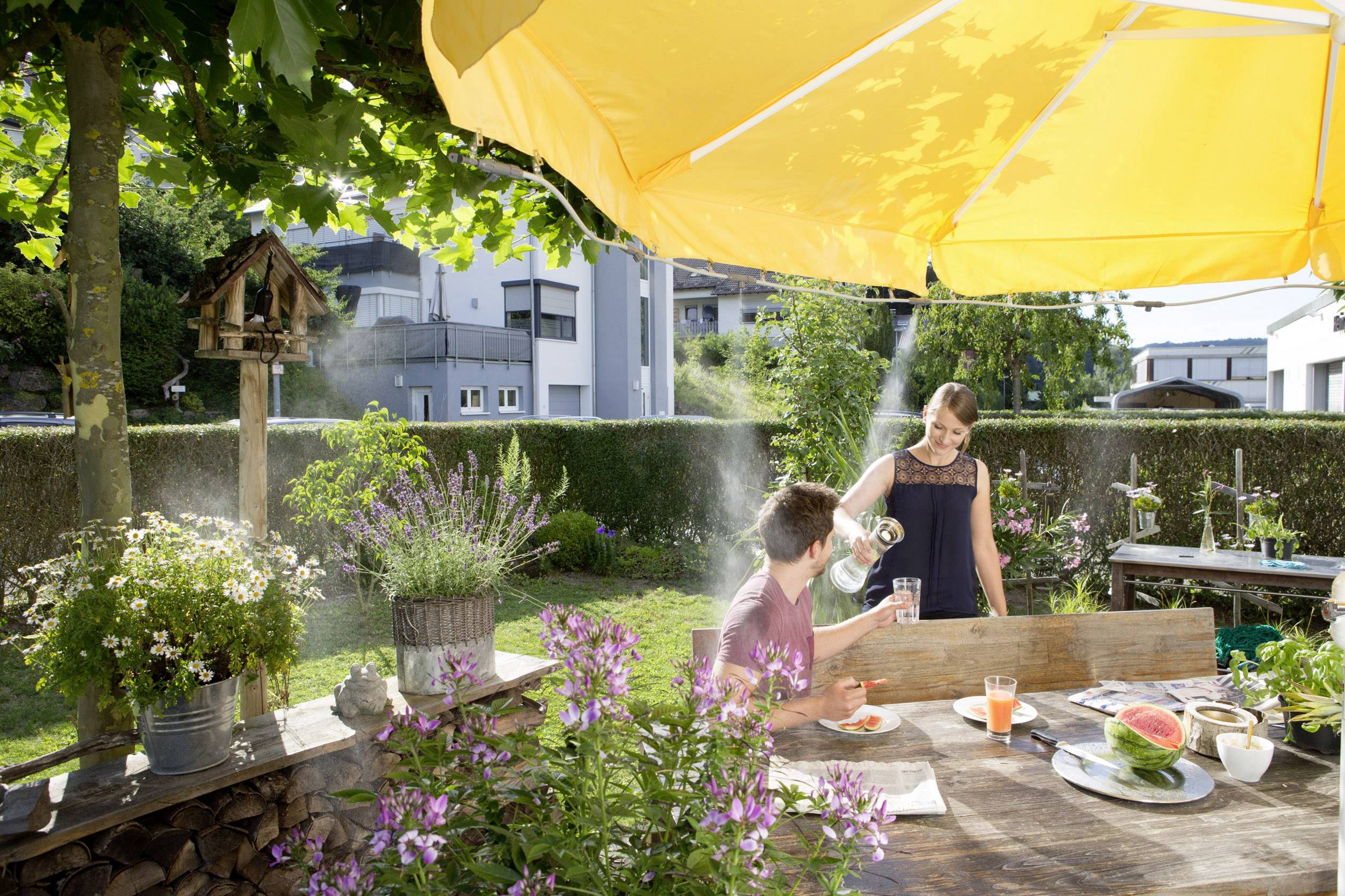 Ein Mann und eine Frau sitzen im sonnigen Garten an einem Holztisch. Er bekommt Wasser eingeschenkt. Um sie herum blühen Pflanzen.