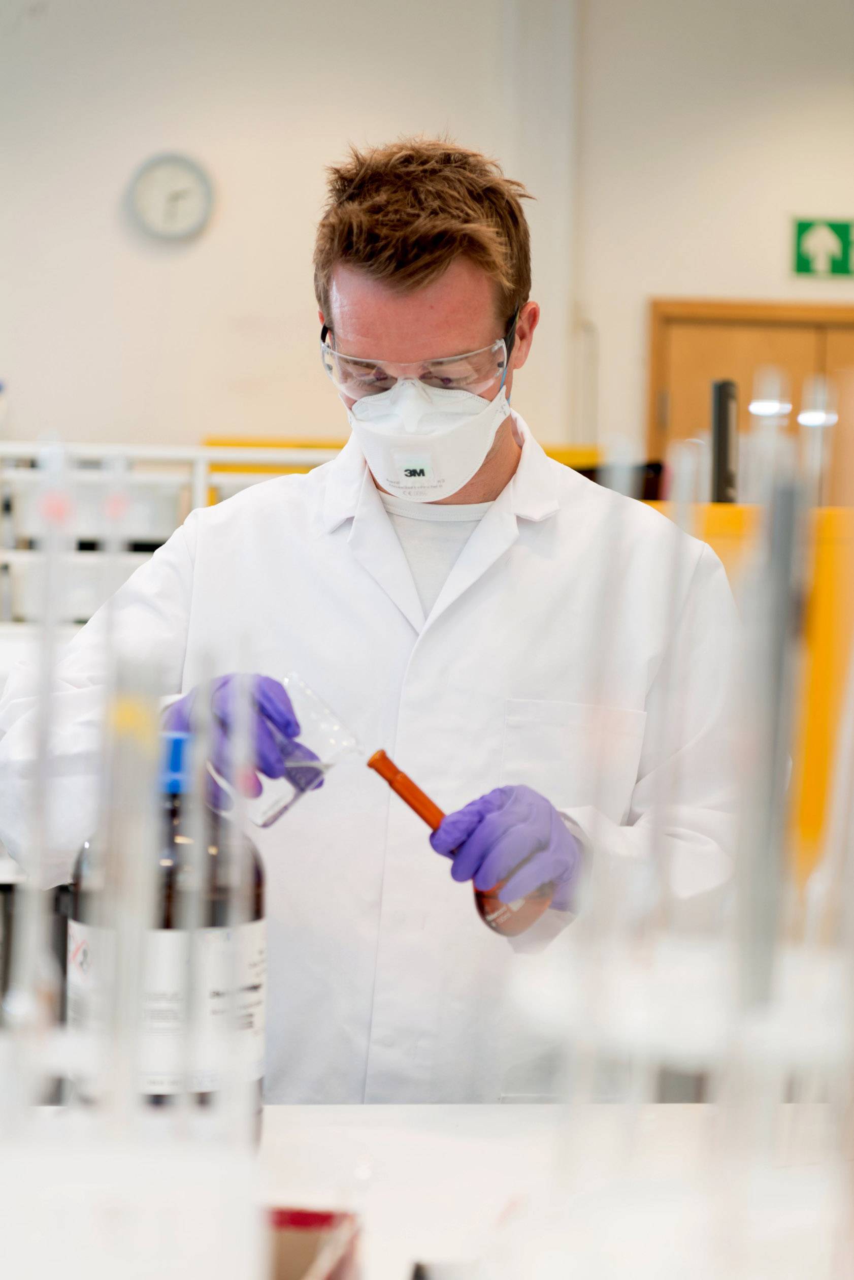 Ein Wissenschaftler im weißen Laborkittel führt in einem Labor mit Schutzbrille und Handschuhen Experimente mit Reagenzglas durch.