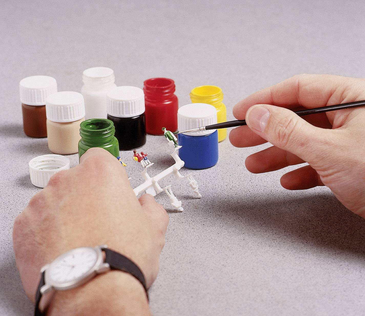 Person malt ein kleines Modell mit einem feinen Pinsel. Im Hintergrund stehen mehrere bunte Farbdosen.