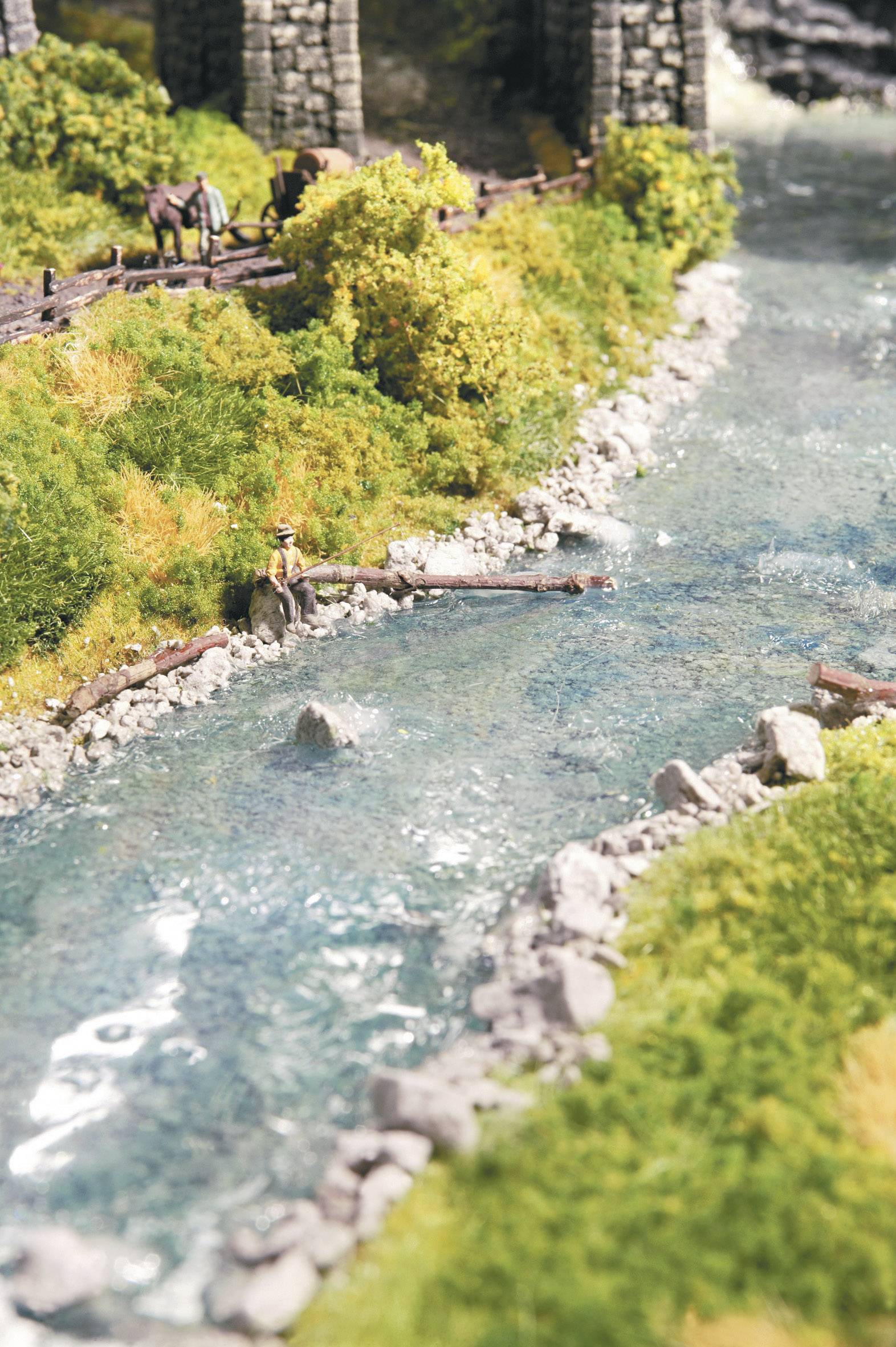 Modelllandschaft mit klarem Fluss, Bäumen, Felsen und einer Figur, die am Ufer sitzt. Pferde stehen in der Ferne nahe einer Holzbrücke.