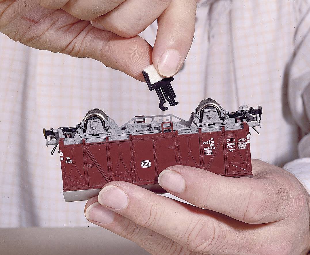 Person hält ein Modellbahnwaggon und befestigt ein kleines Bauteil am Boden. Hände fixieren den Waggon, um das Anbringen zu erleichtern.