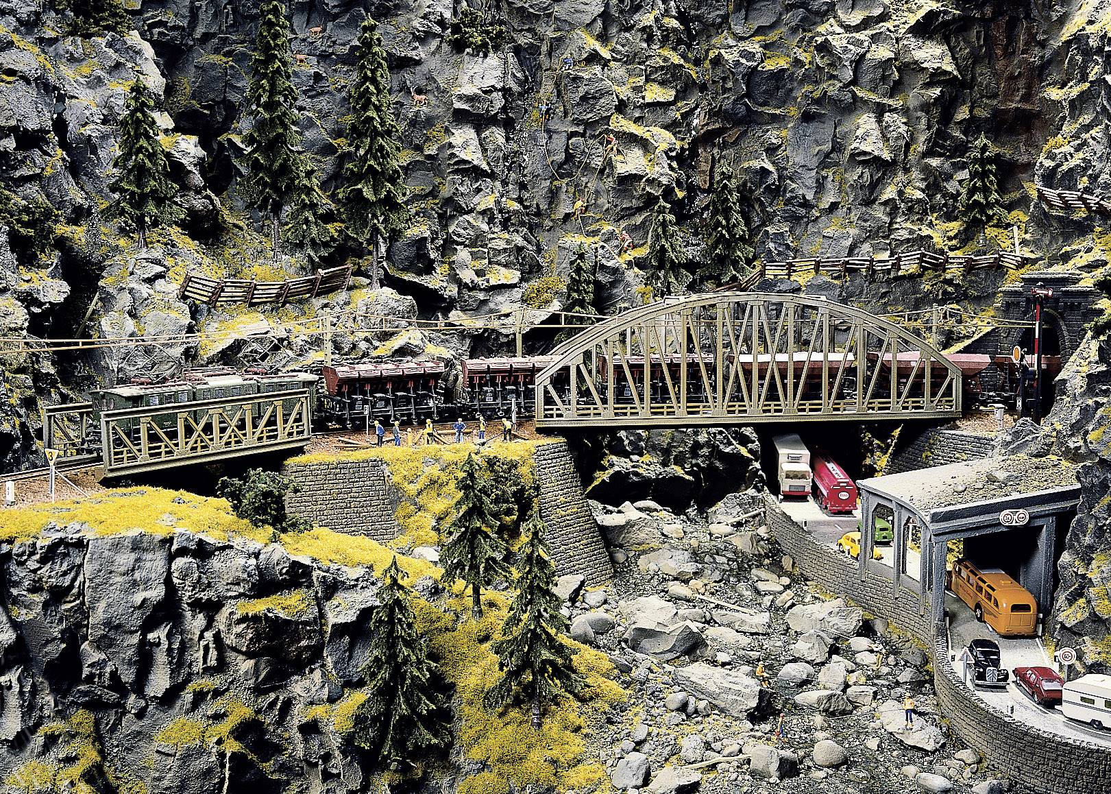 Eine detailreiche Modelleisenbahnanlage zeigt Züge auf Brücken und durch Tunnel in einer felsigen, bewaldeten Landschaft.