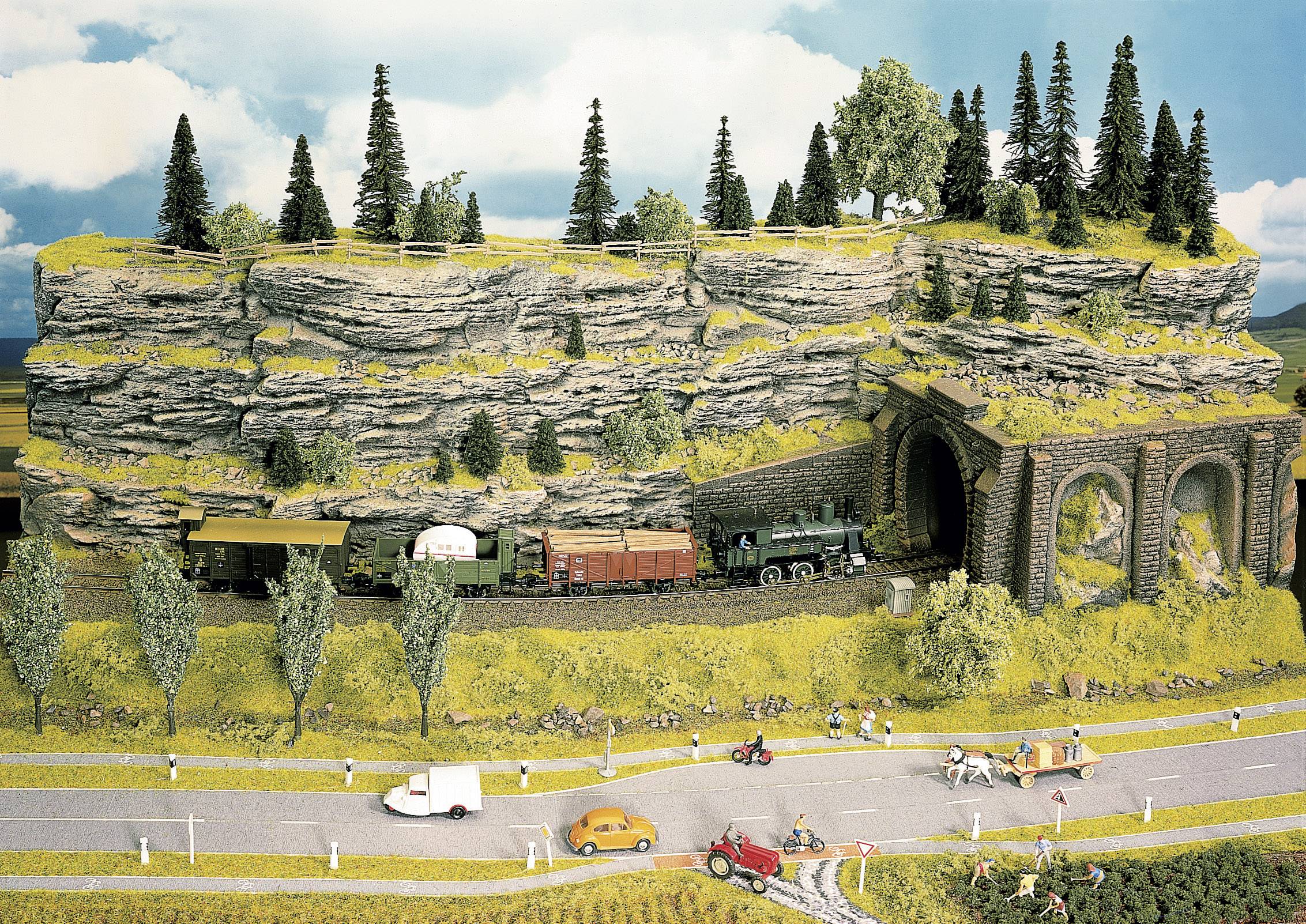 Modelleisenbahn-Diorama mit einem Zug, der durch einen Tunnel in einer felsigen, bewaldeten Landschaft fährt. Straße im Vordergrund.