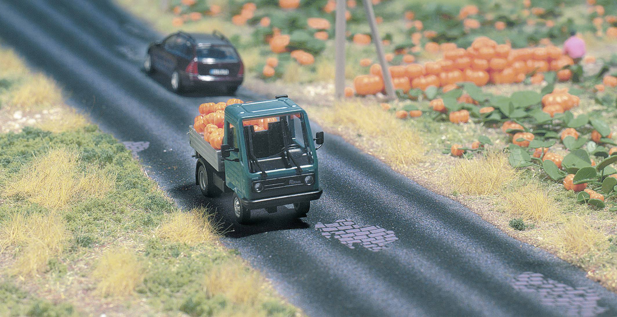 Ein kleines Modell-Lkw transportiert Kürbisse auf einer Landstraße, gefolgt von einem Modellauto. Umgebung mit Feldern und Kürbissen.